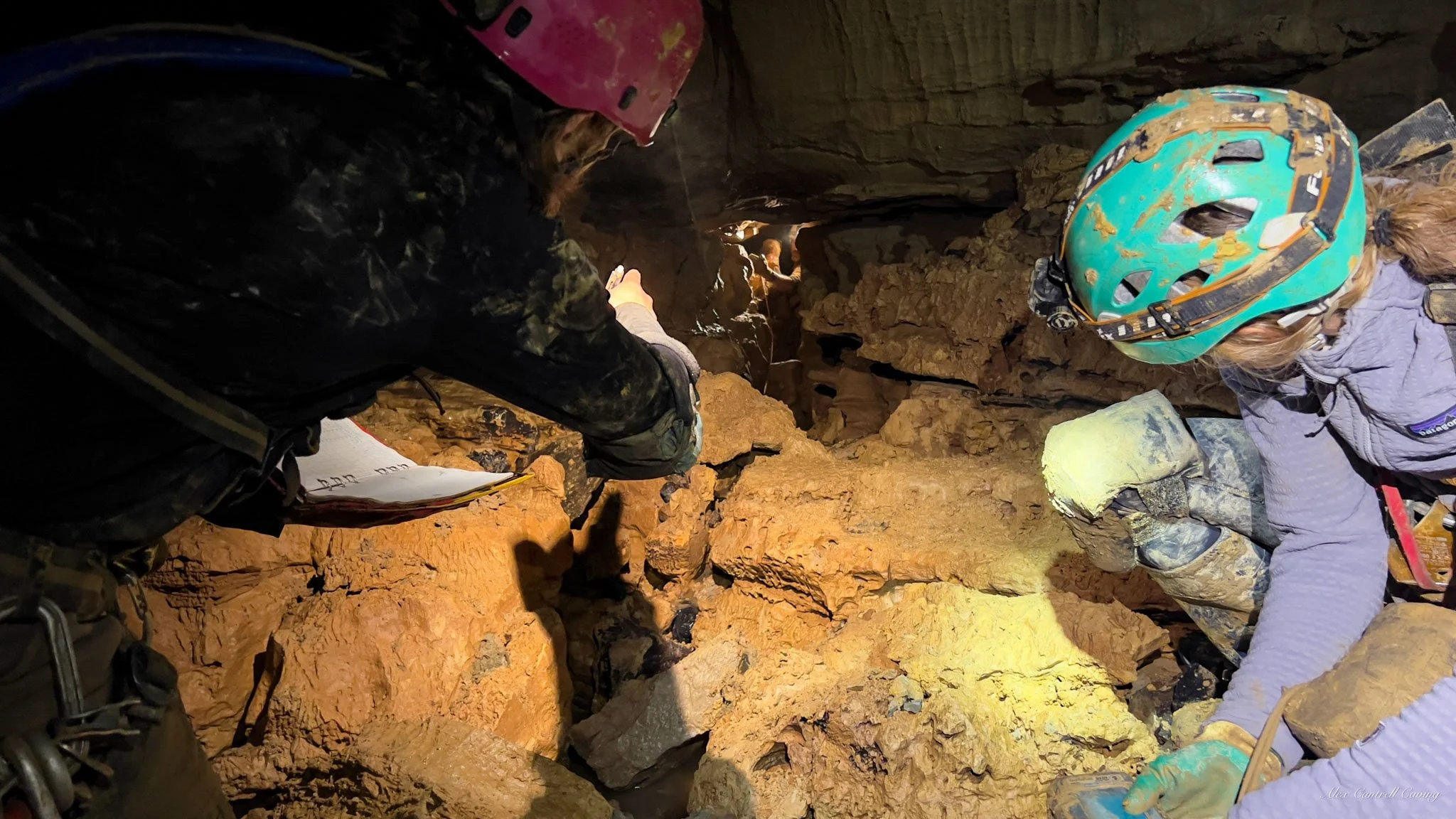 Two people wearing helmets and headlamps exploring a cave with rocky walls.