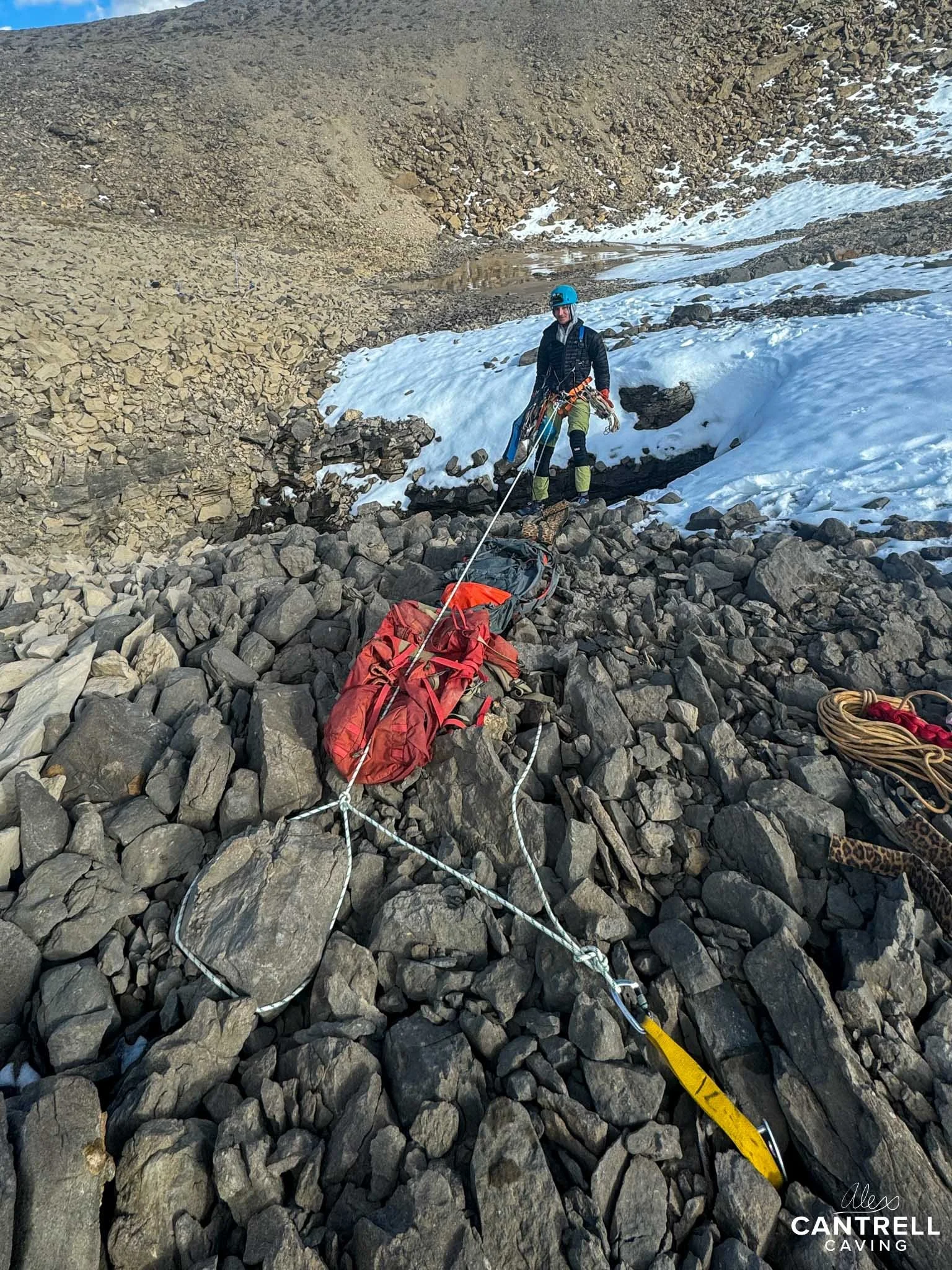 Person caving on rocky terrain with safety gear, ropes, and a red bag, in a snowy mountainous area.