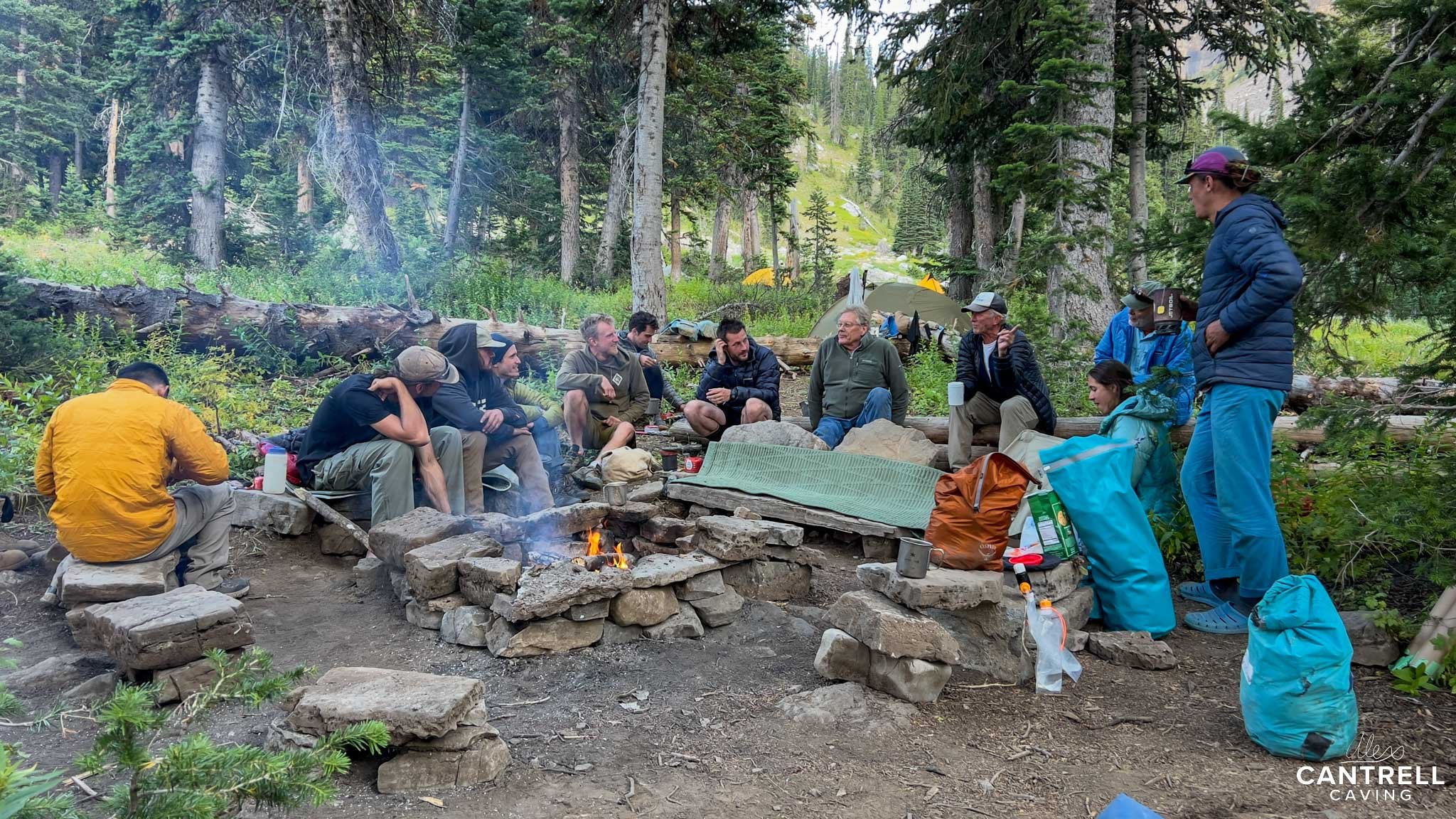 A group of campers sitting around a stone fire pit in a forest clearing, with tents visible in the background. Various camping gear is scattered around, and the campers are dressed in outdoor clothing.
