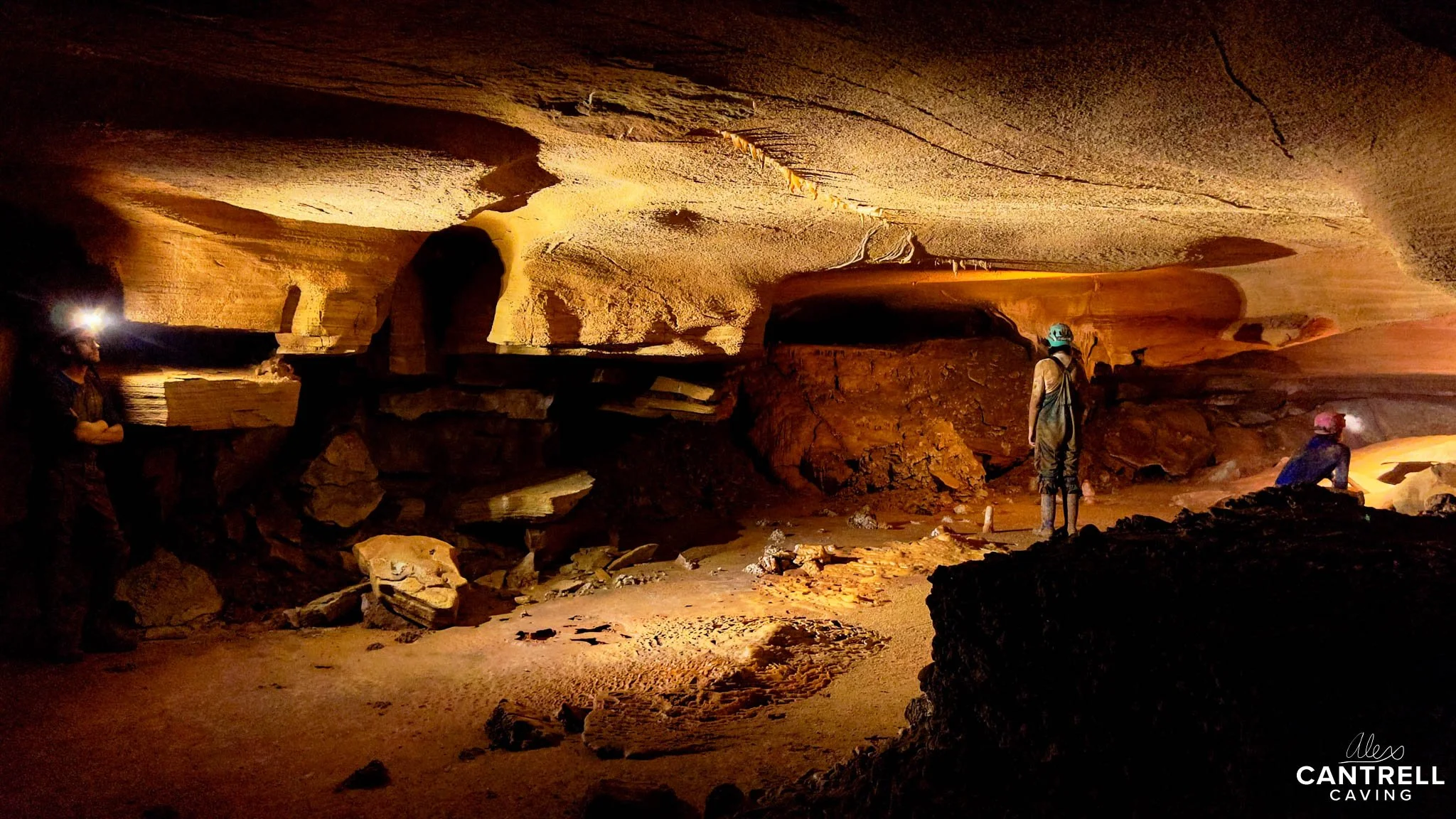 Cave interior with rough rock formations and explorers wearing helmets and headlamps.