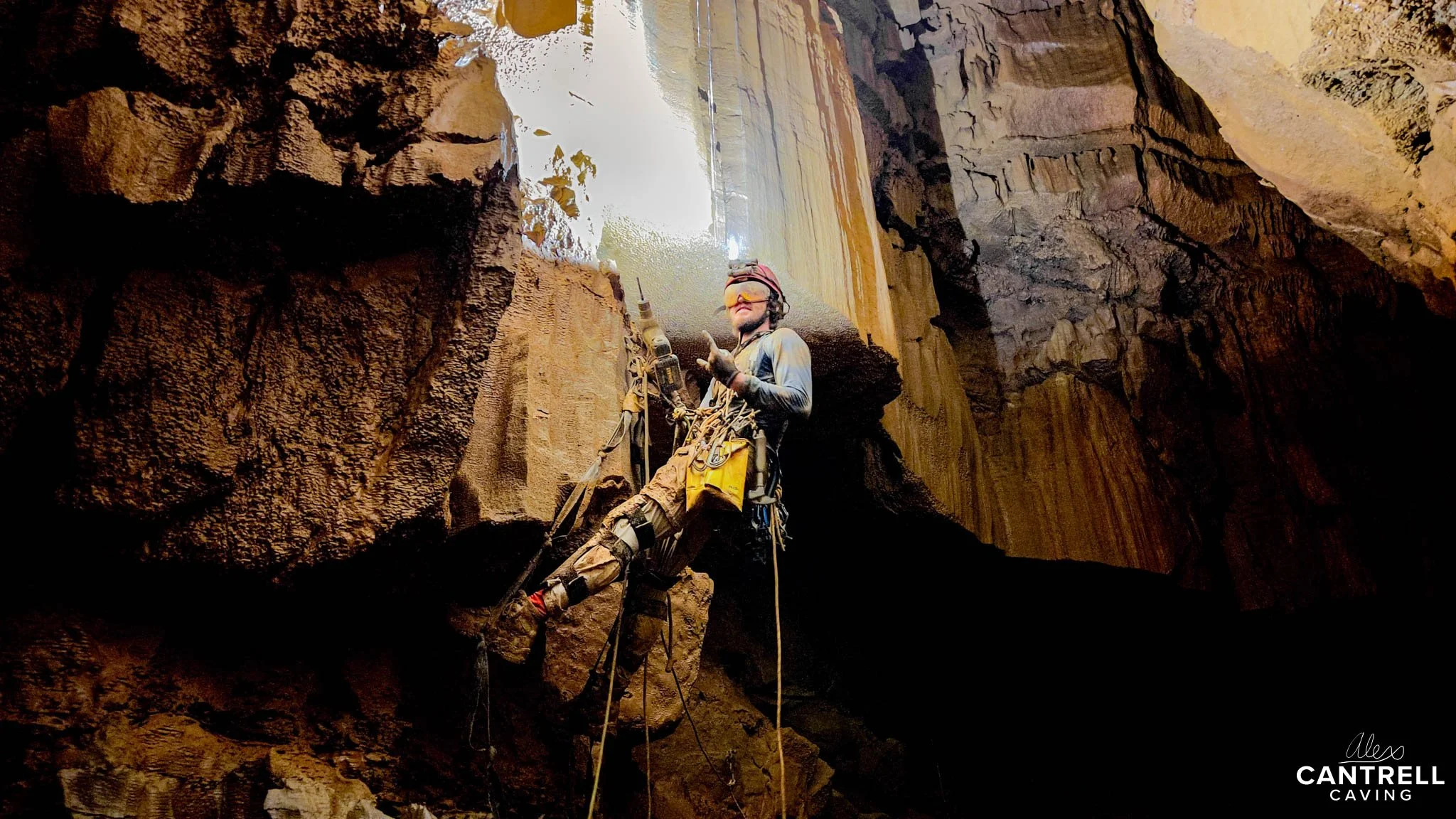 Caver climbing in a cave with harness and ropes, illuminated shaft of light above.