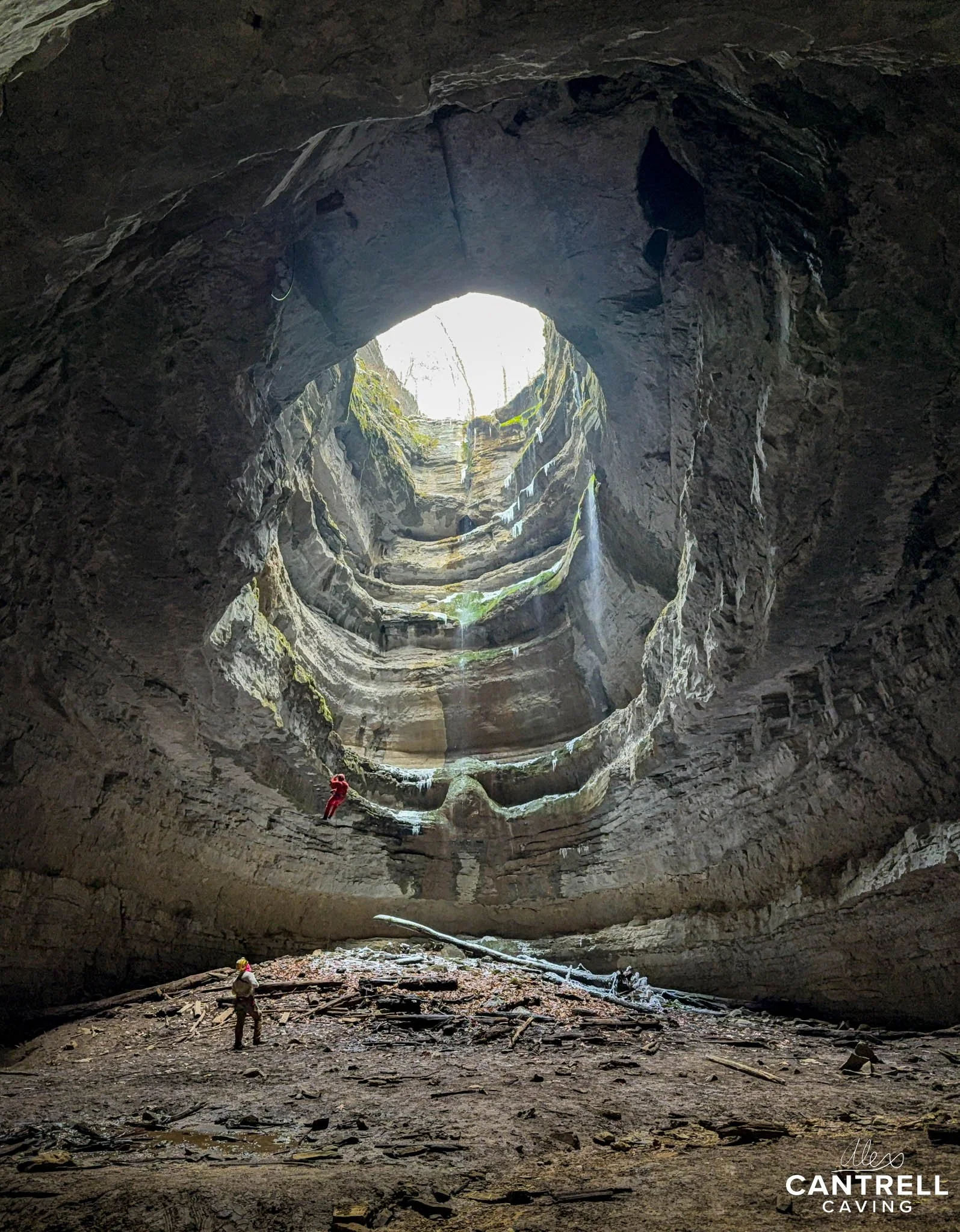 Cave with a large vertical opening, featuring a person in red gear rappelling down and another standing on the ground. Light streams through the opening, illuminating the rocky walls and debris below.