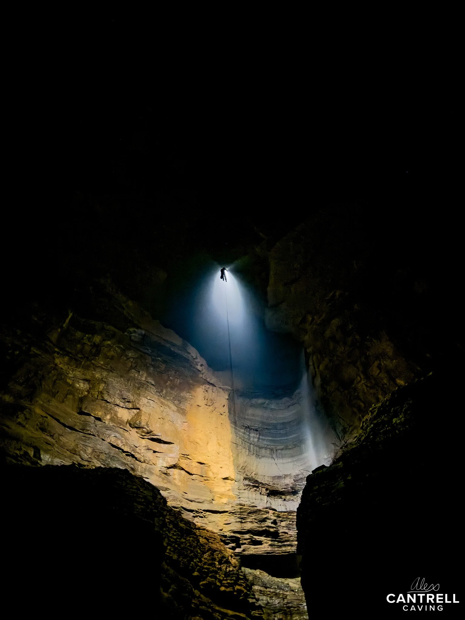 Silhouette of a person rappelling inside a large cavern with light streaming in from above, illuminating the rock walls.