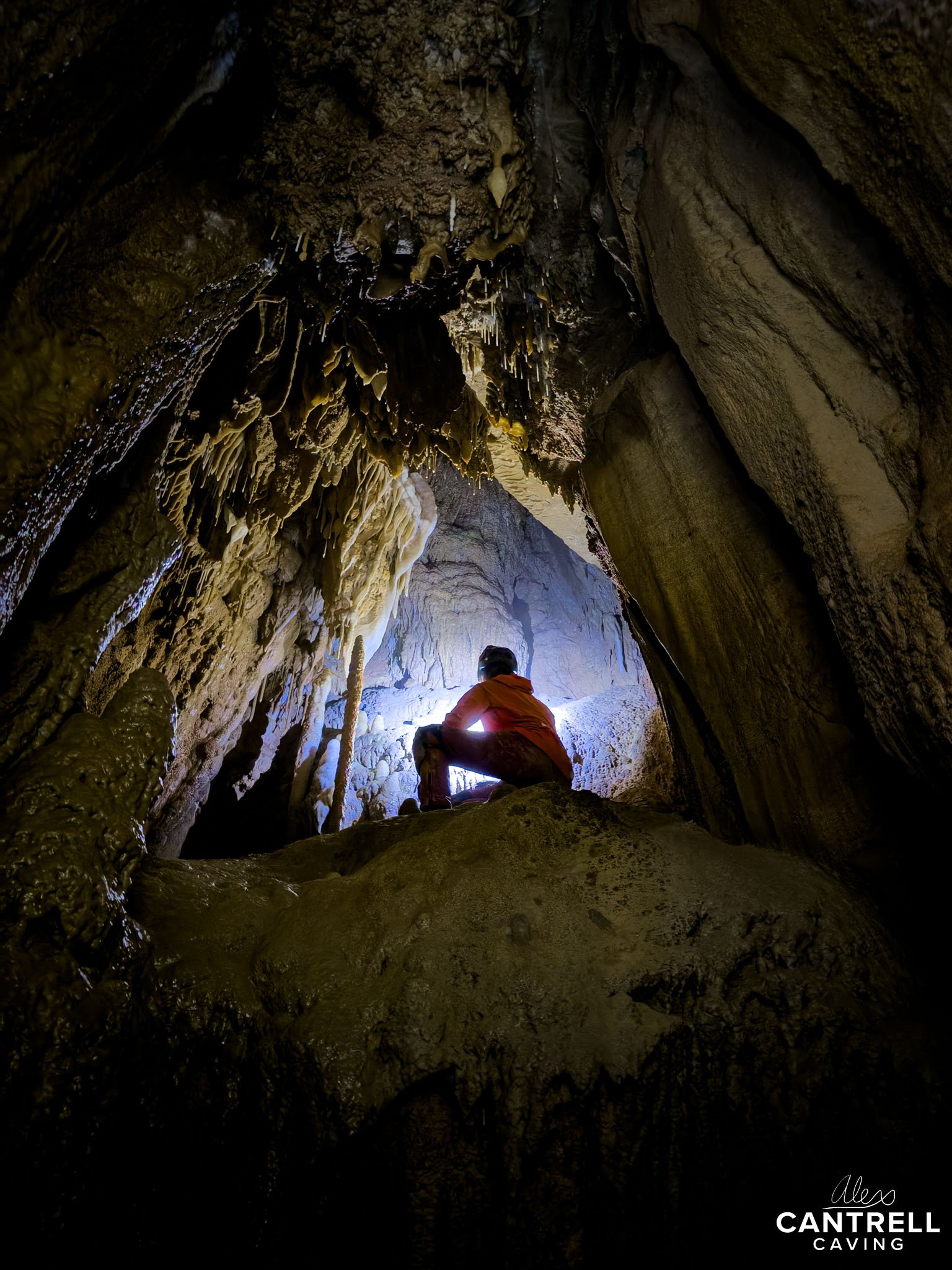 Person wearing a helmet and headlamp exploring a dark cave with stalactites and rock formations.