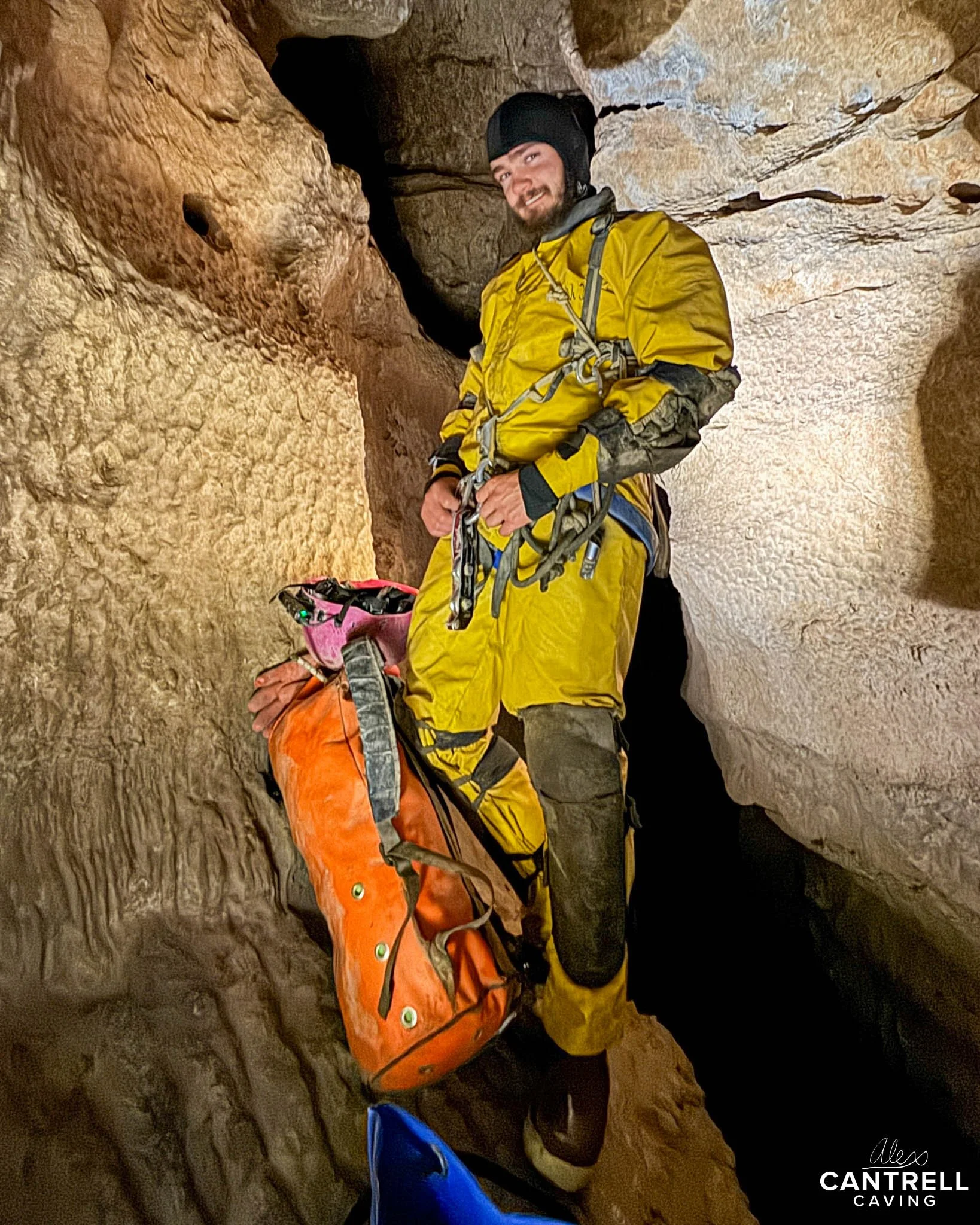 Person wearing yellow caving suit and helmet inside a cave, with ropes and gear, standing next to an orange bag.