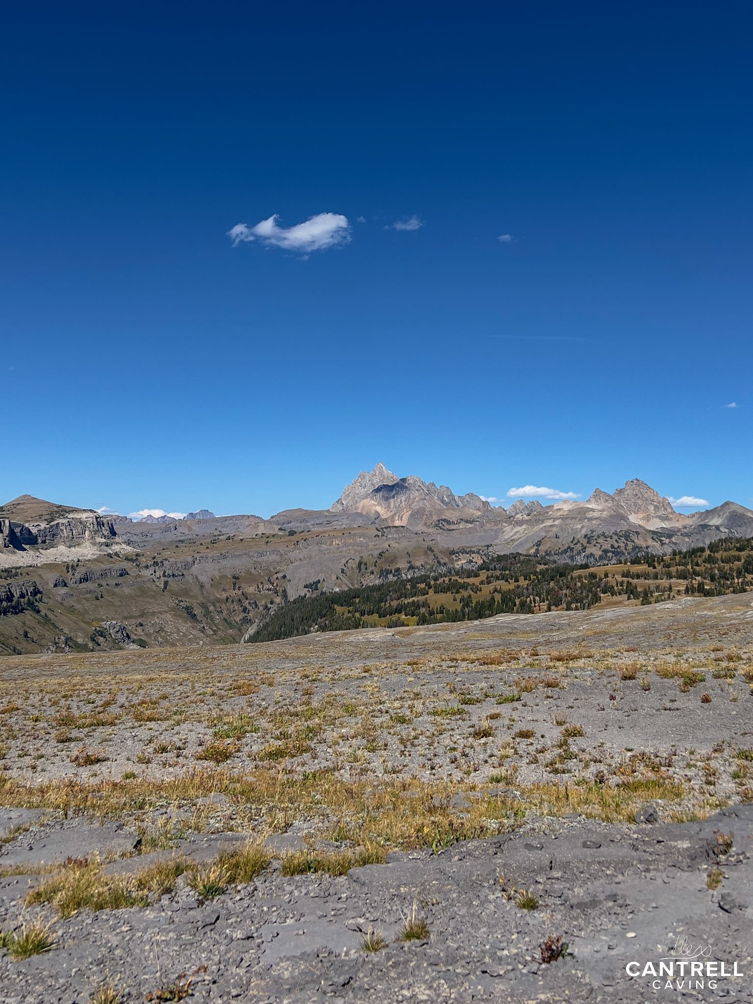 Rocky mountain landscape with distant peaks and a clear blue sky. Sparse vegetation covers the foreground.