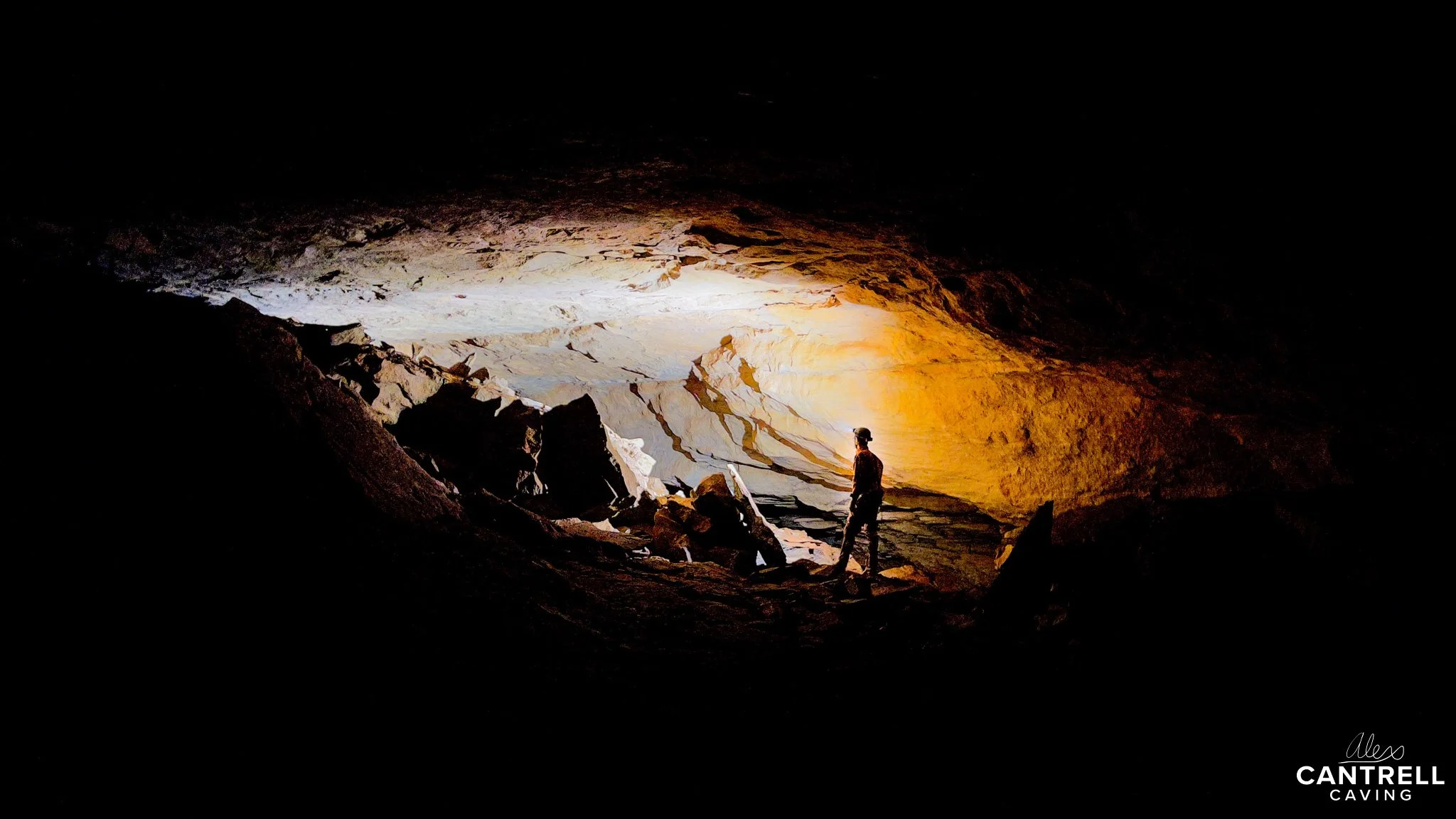 Cave interior with explorer in silhouette standing near rocky formations, illuminated by artificial light.