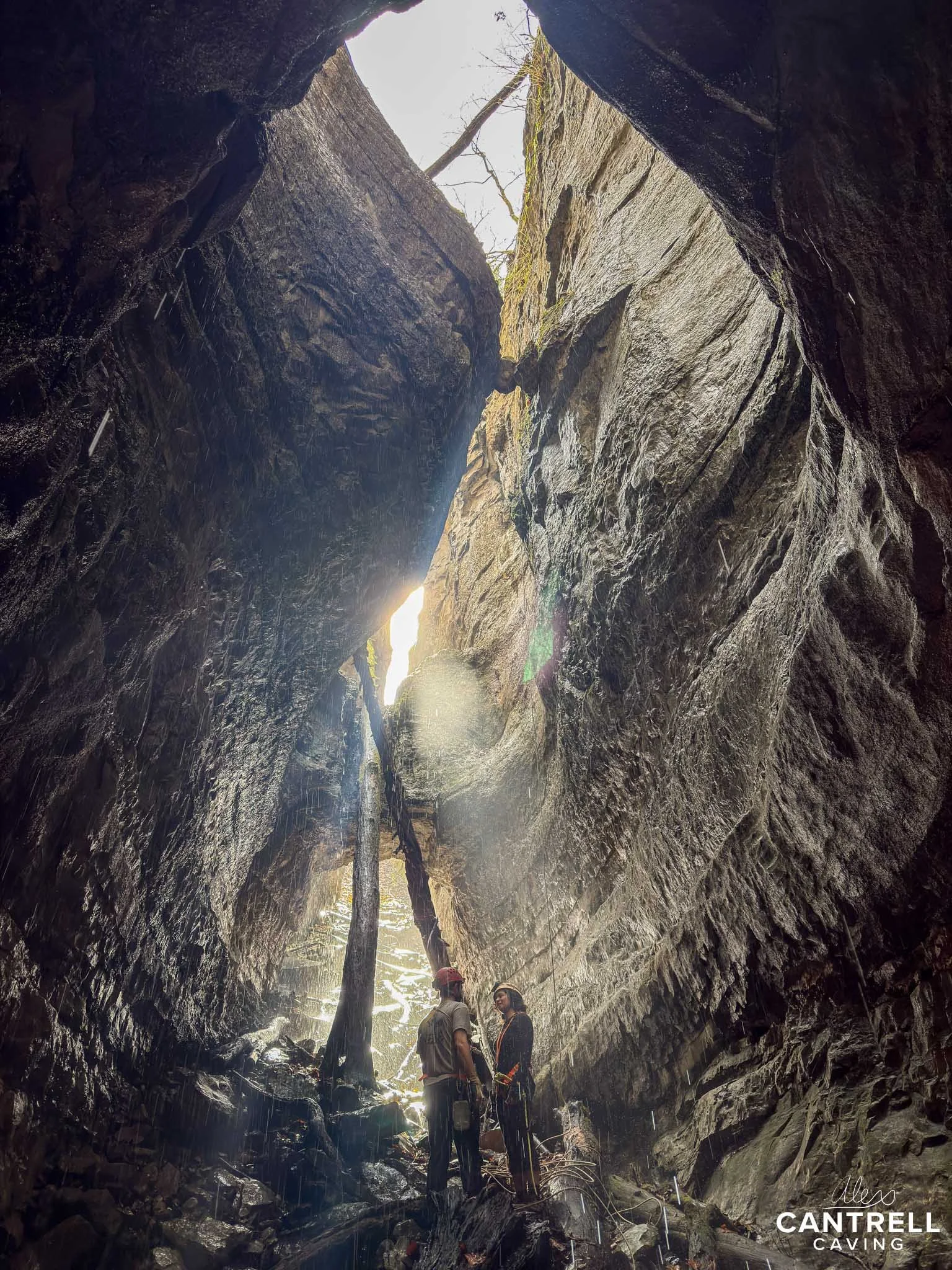 Two people wearing helmets stand inside a large, dimly lit cave with steep rock walls and natural light shining through an opening at the top. The ground is rocky and wet, with a tree trunk leaning inside the cave. The scene is dramatic and adventuro