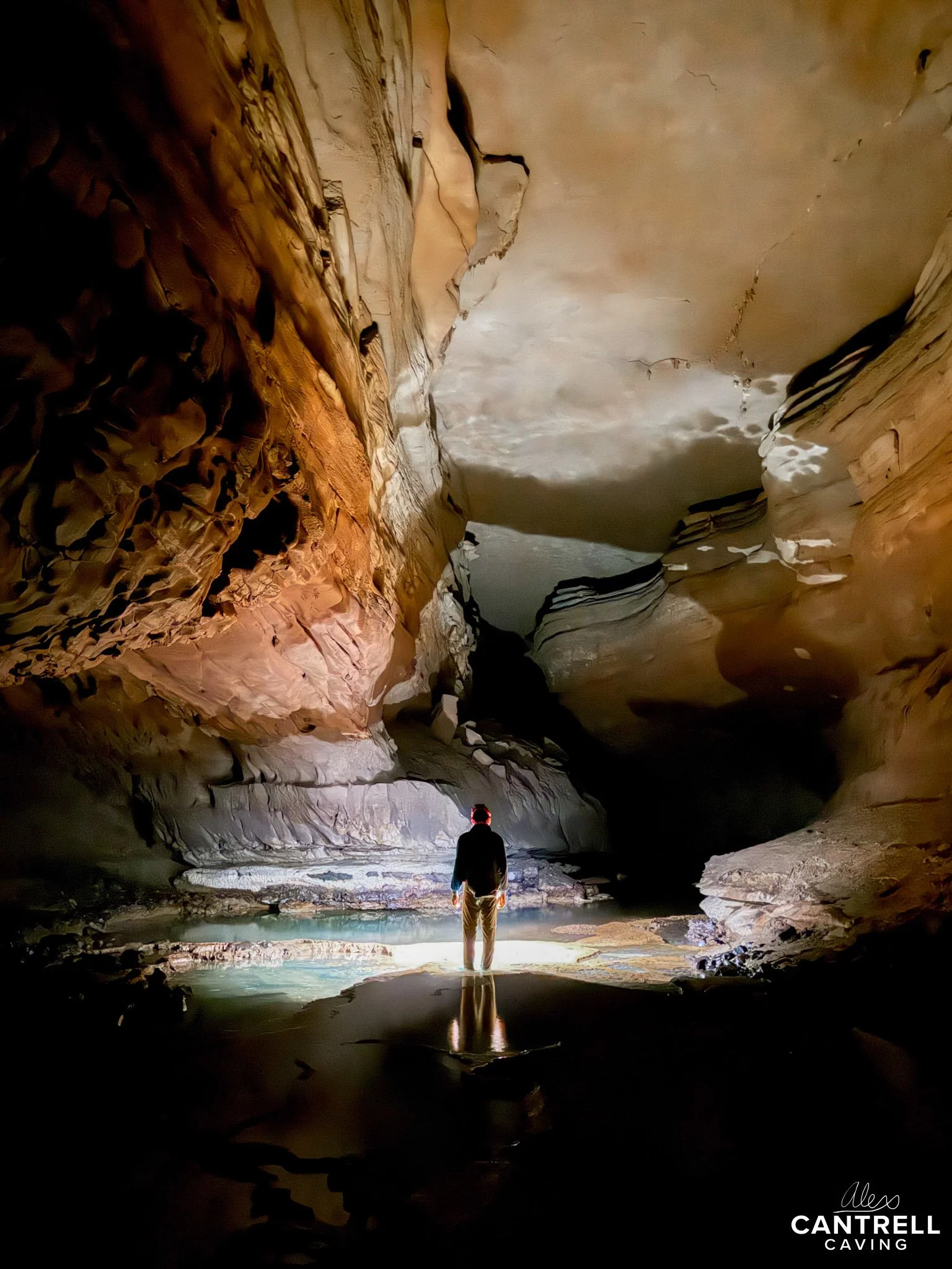 Person standing in a dimly lit cave, illuminated by a light source reflecting on the water, surrounded by rocky walls and formations.