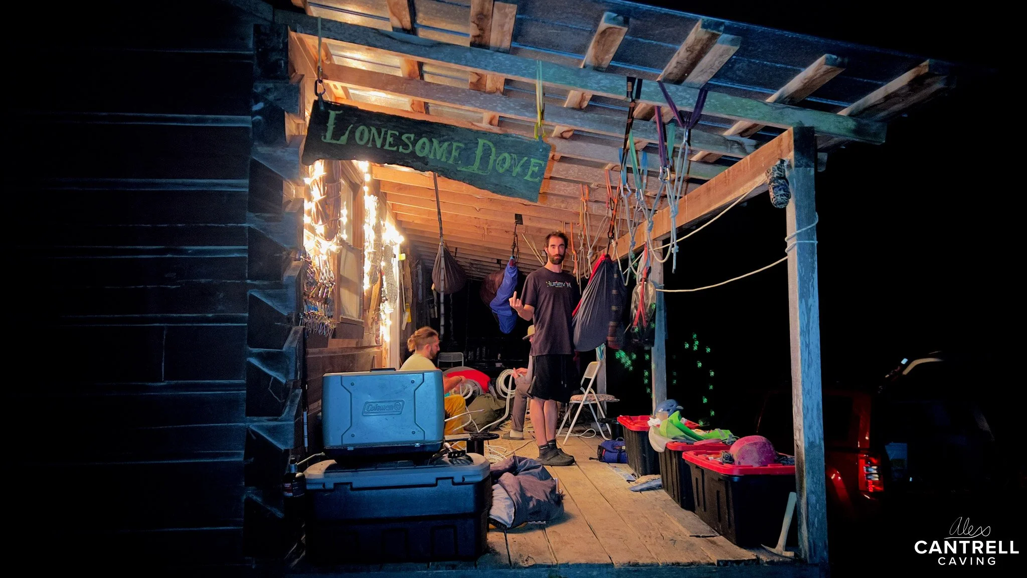 Outdoor porch at night with two people, gear storage, string lights, and a "Lonesome Dove" sign.