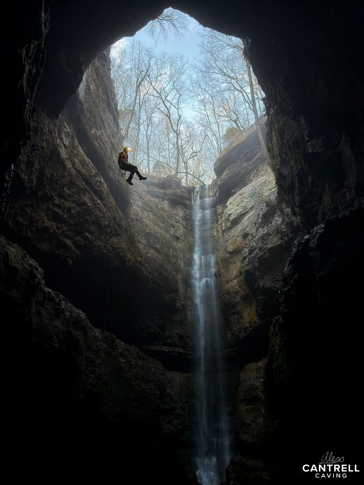 Person in caving gear rappelling down inside a large cave with a waterfall and view of sky and trees through the cave opening.