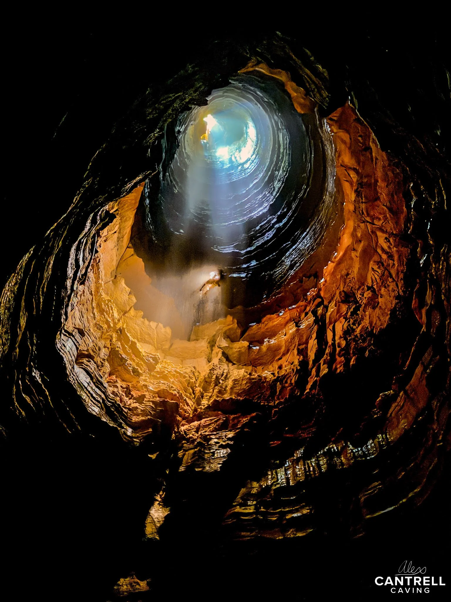 Person illuminated in a deep cave with sunlight streaming from an opening above and rugged rock walls surrounding them.