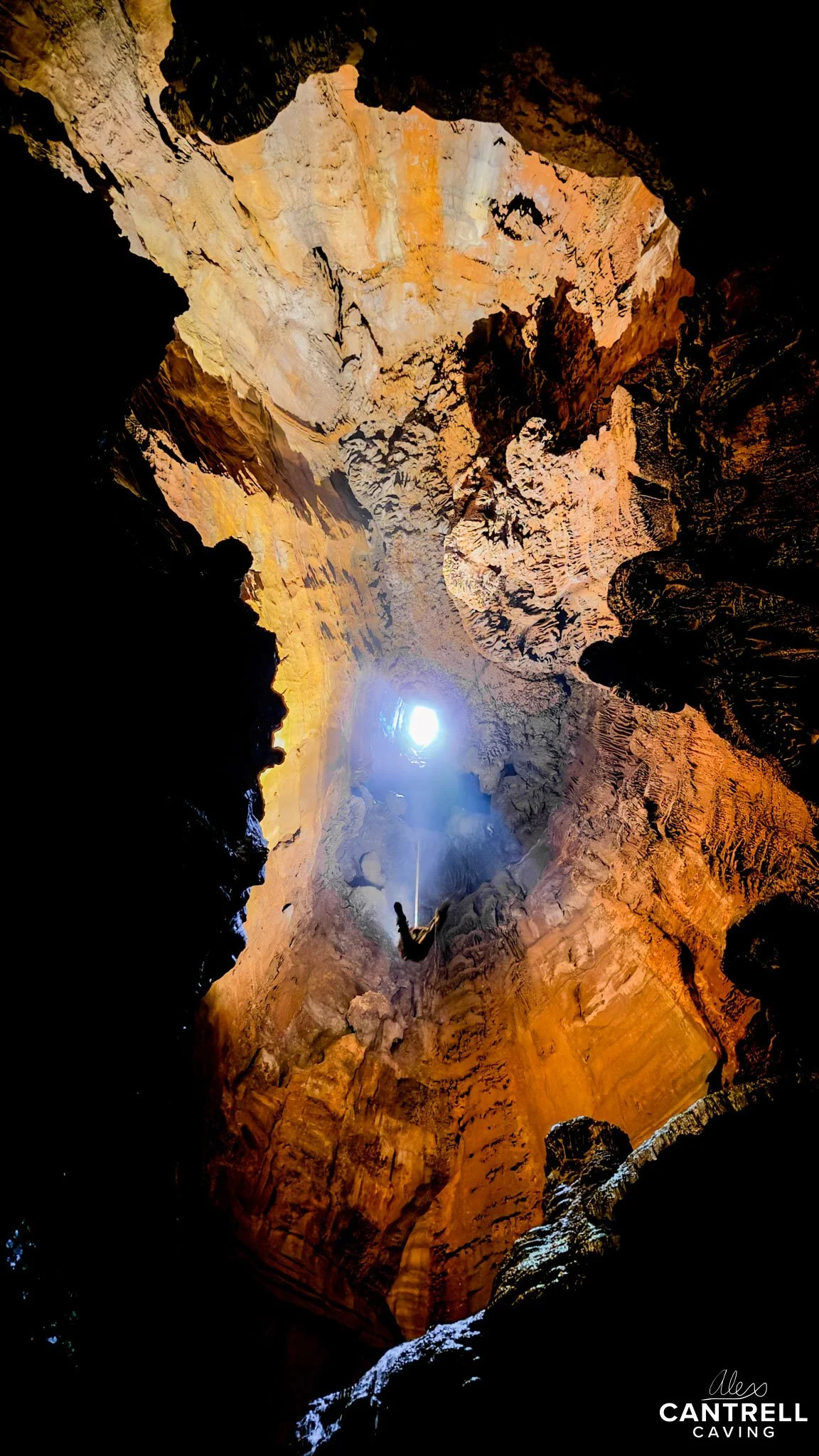 Person rappelling down a large cave with rugged rock walls and sunlight shining through the opening at the top.