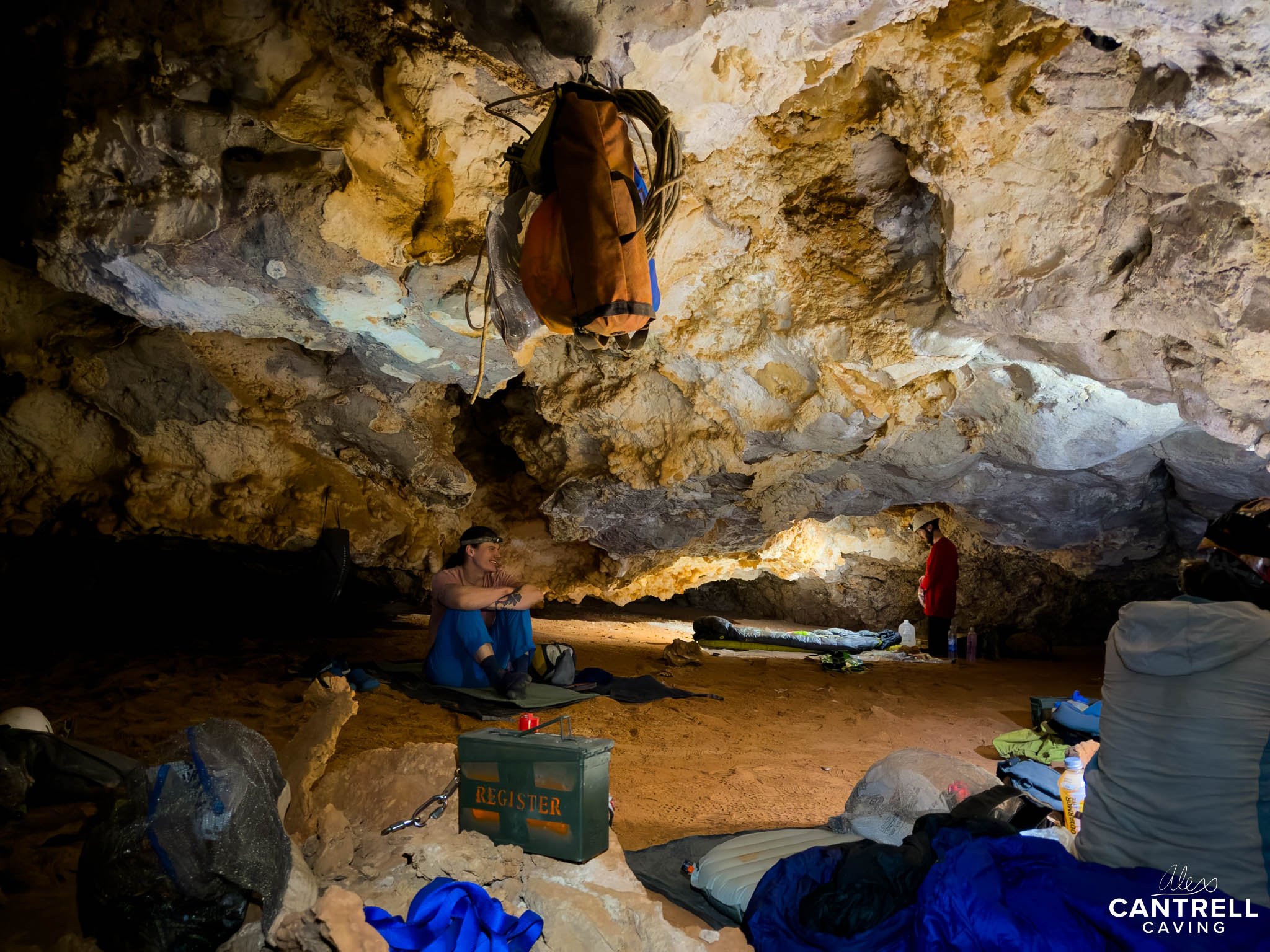 Interior of a cave with cavers preparing gear; one person sitting on a mat, surrounded by camping supplies, ropes, and bags, with rocky cave walls in the background.