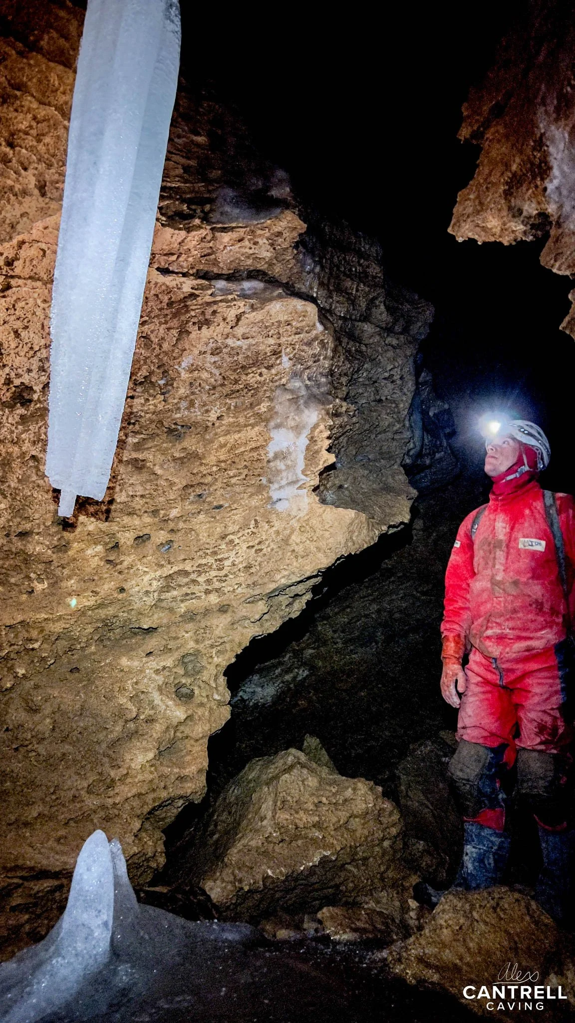 Caver wearing a helmet with headlamp exploring a cave with large icicles hanging from the ceiling.