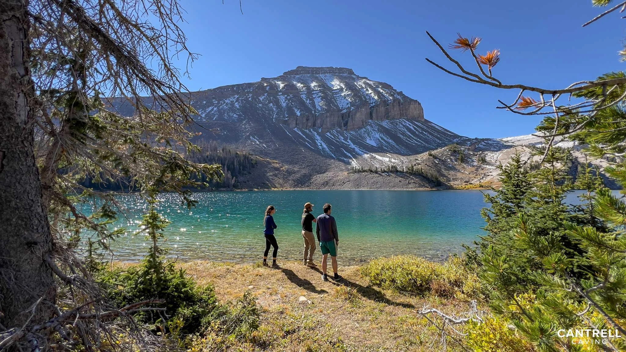 Three people standing by a lake with a mountain in the background, surrounded by trees and clear blue sky.