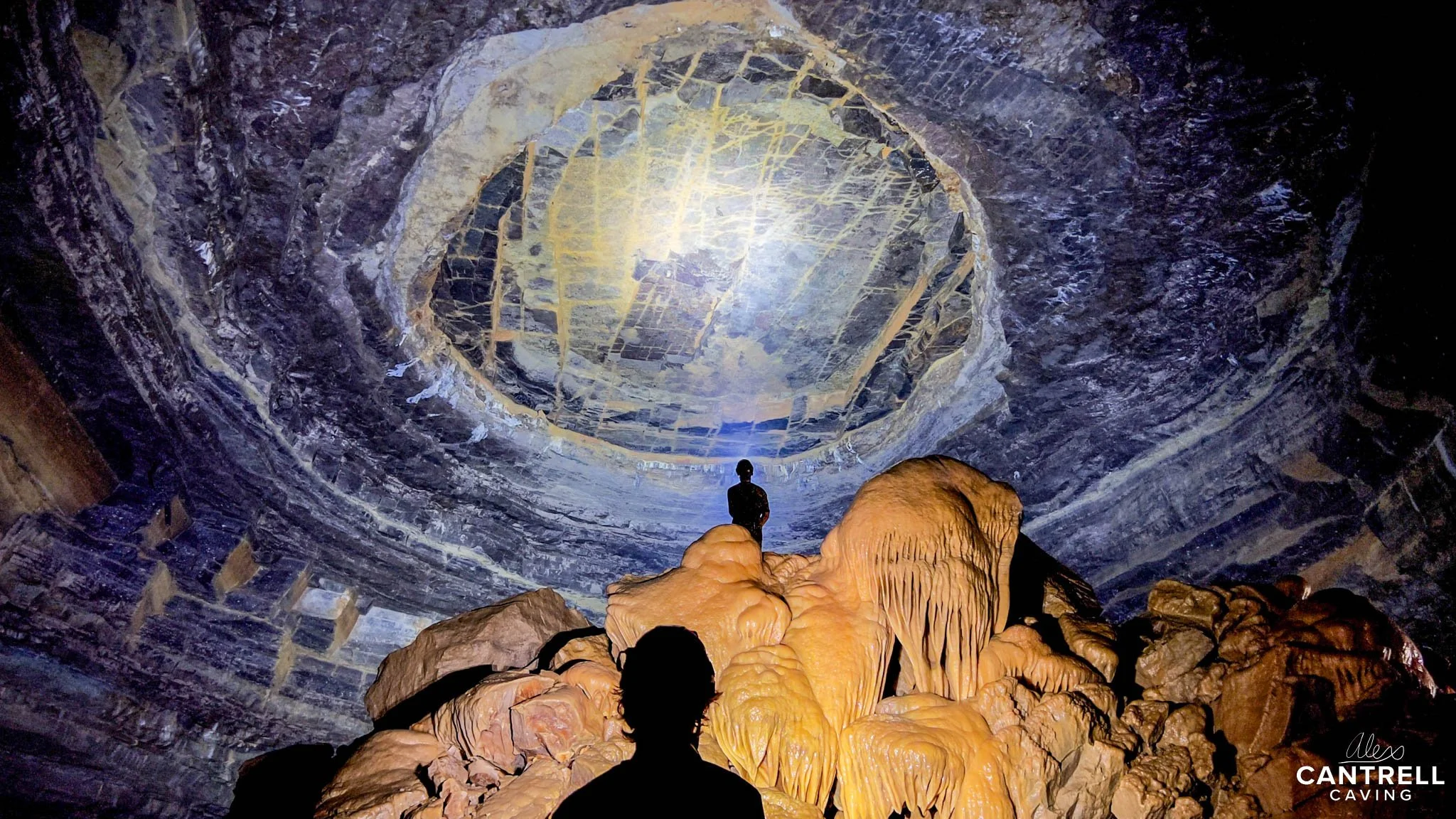 Two people exploring a large cave chamber with stalactites and a textured ceiling, illuminated by artificial light. The cave walls are dark with orange formations in the foreground.