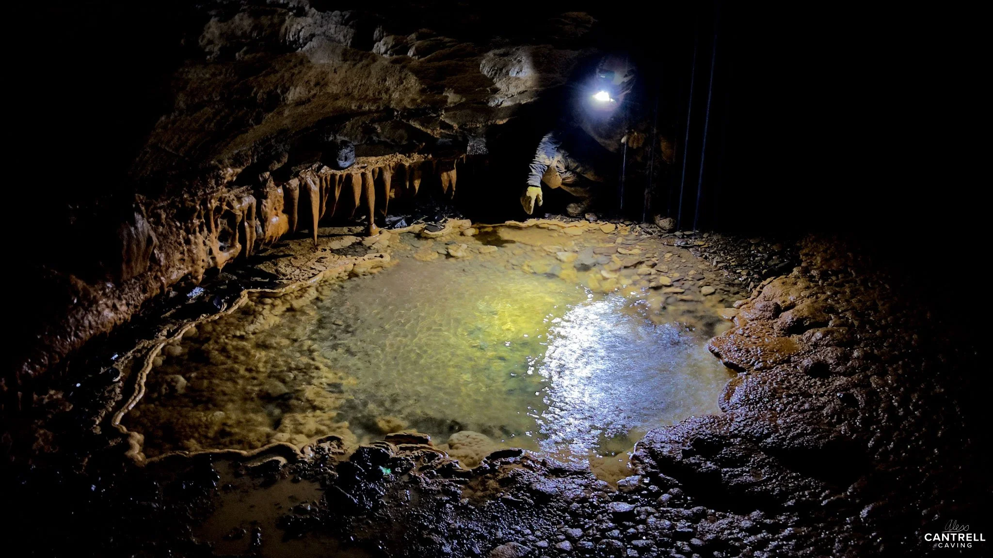 A dark cave interior with a natural pool of water and rock formations, illuminated by a bright light held by a person wearing caving gear.