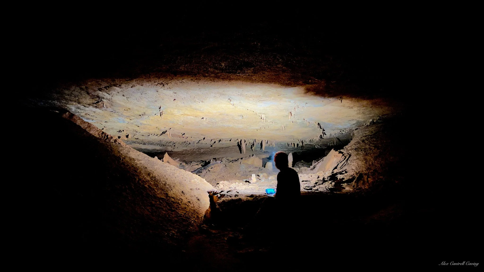Silhouette of a person sitting inside a dimly lit cave, with stalactites hanging from the ceiling.