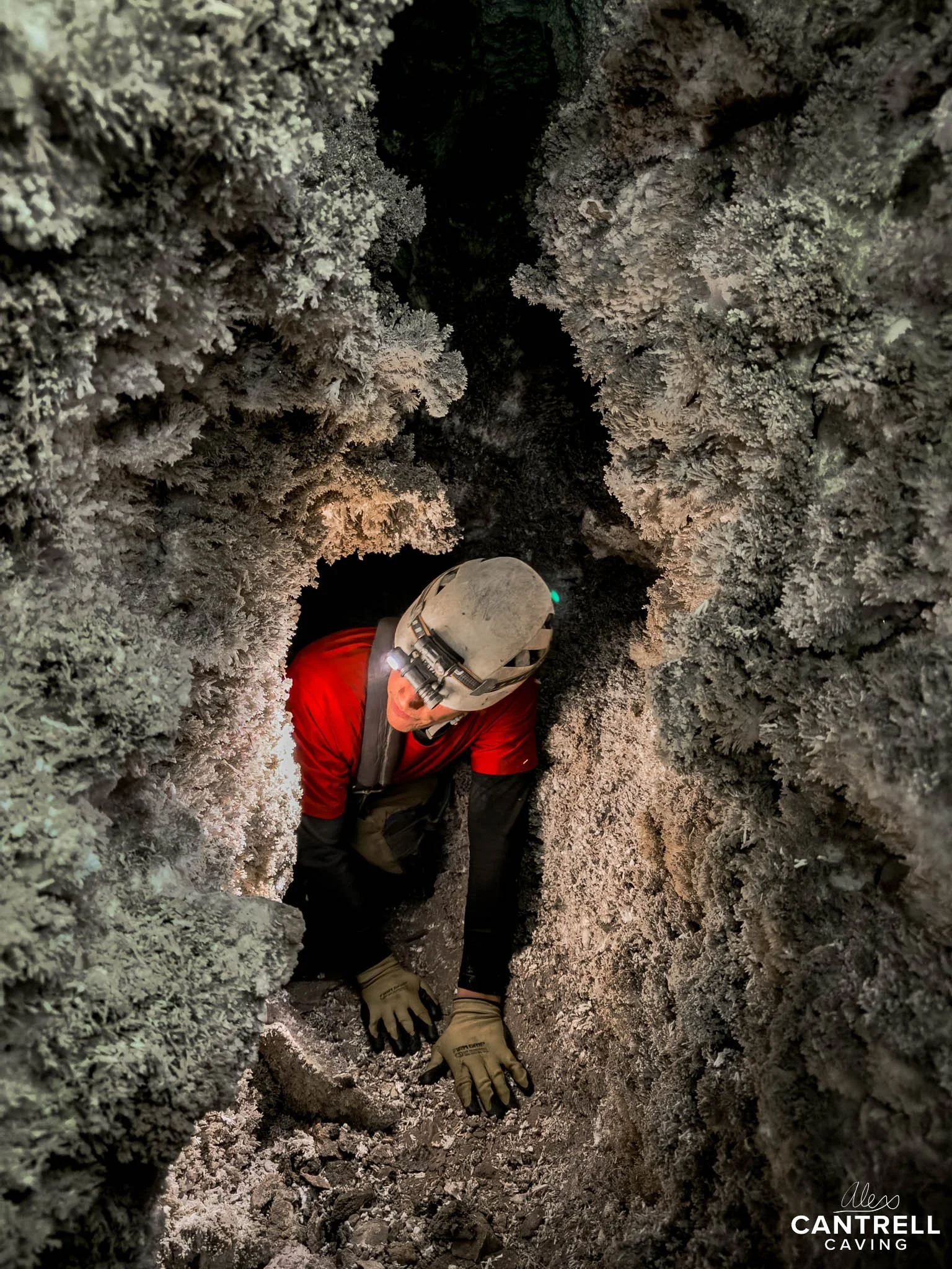 Person wearing a helmet and gloves exploring a narrow cave passage with rocky and crystalline formations.