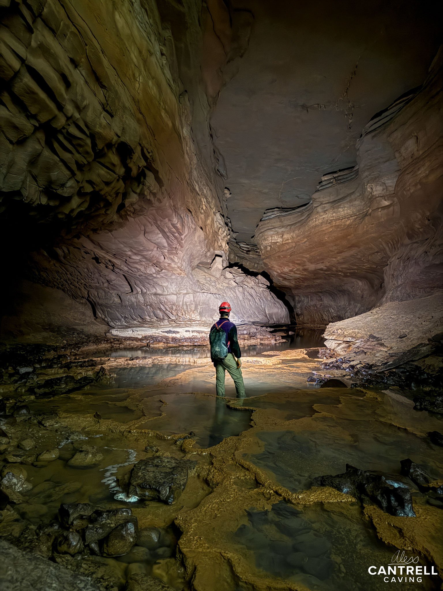A person wearing a caving helmet and backpack exploring a large underground cave with rocky walls and formations. The cave floor is partially submerged in water.