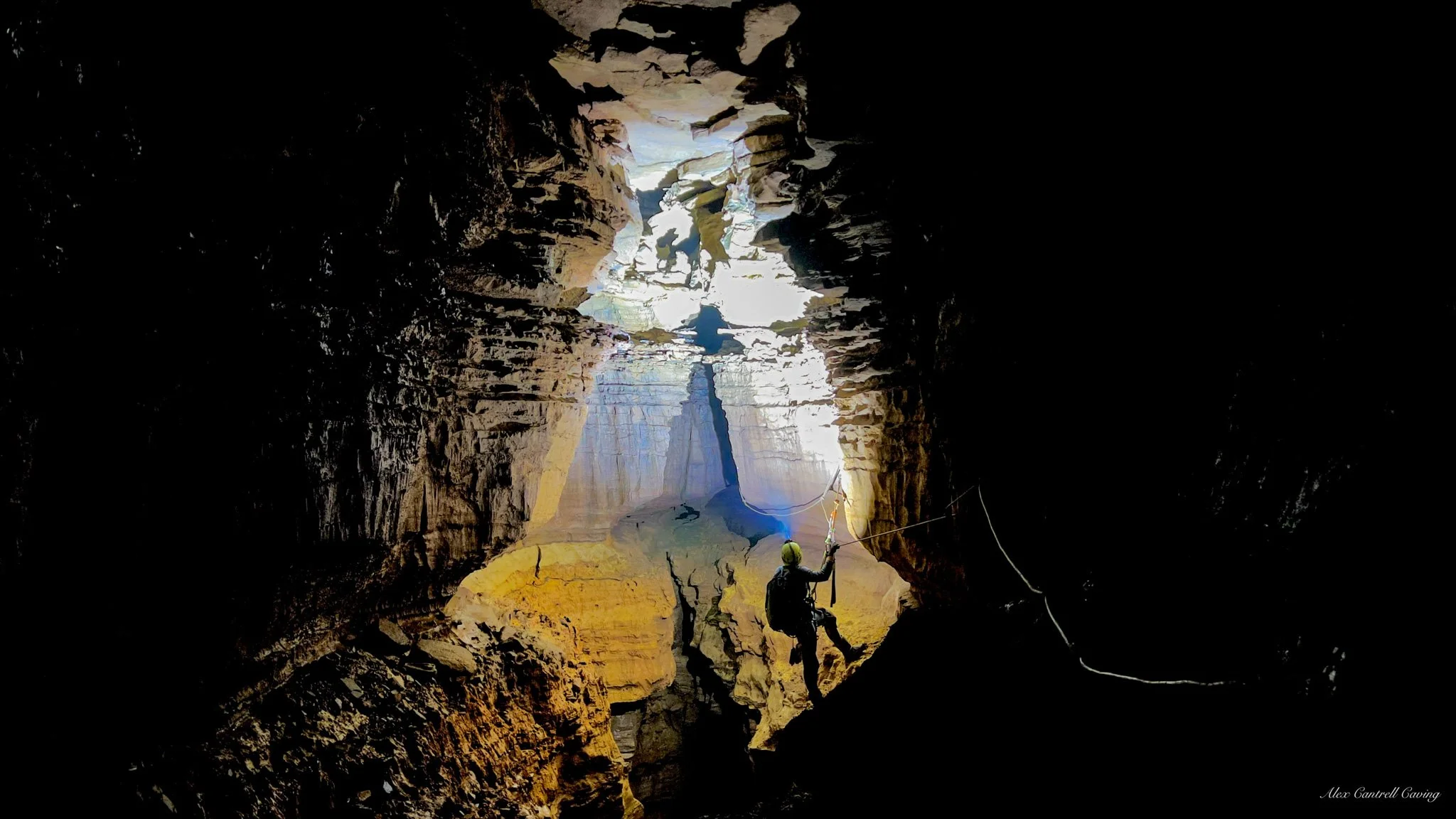 Cave explorer rappelling into a deep, illuminated cavern with rocky walls.
