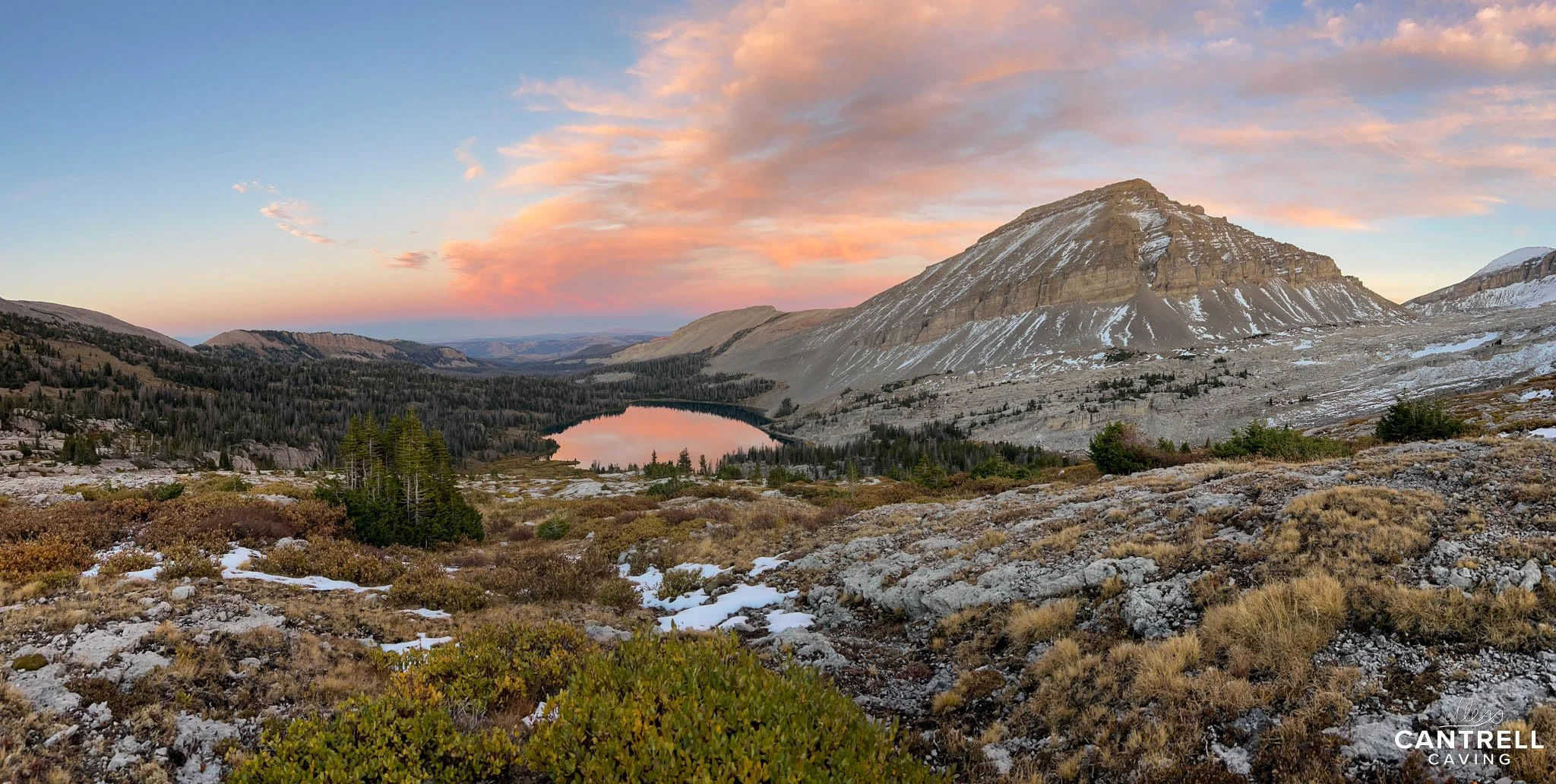 Mountain landscape at sunset with snow patches, a lake reflecting the sky, and rugged terrain with scattered trees.