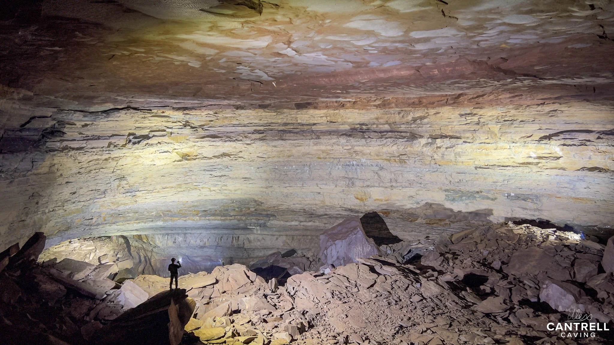 Wide view of a large cave interior with a person standing on rocky terrain. The cave walls show layers of textured rock, illuminated by lights. The ceiling has a mix of colors ranging from brown to beige.
