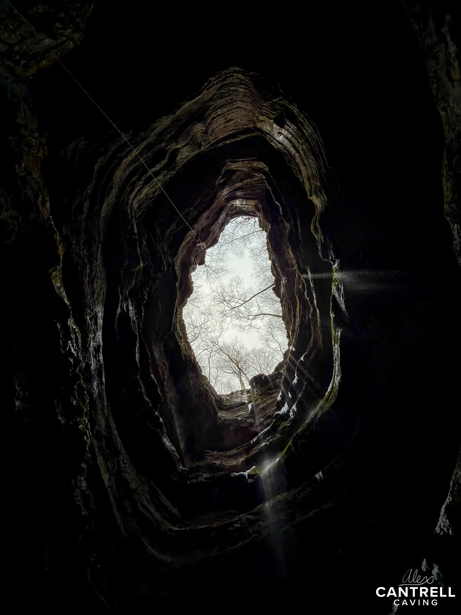 View from the bottom of a vertical cave shaft looking up towards the sky, with rock layers surrounding the opening and some sunlight filtering through.