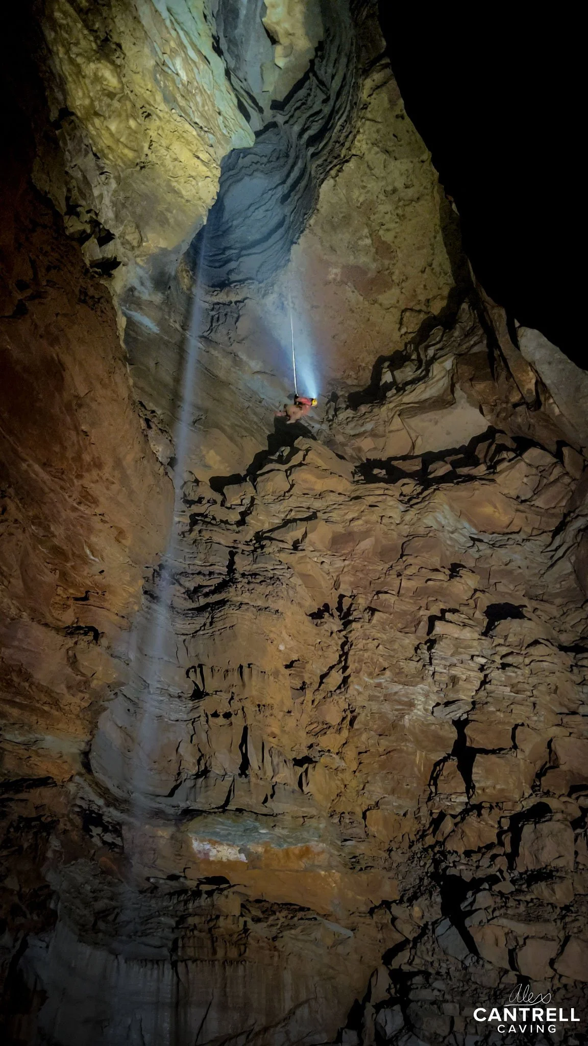 Person wearing a helmet rappelling inside a large, rocky cave, with light beams highlighting the rugged surfaces.