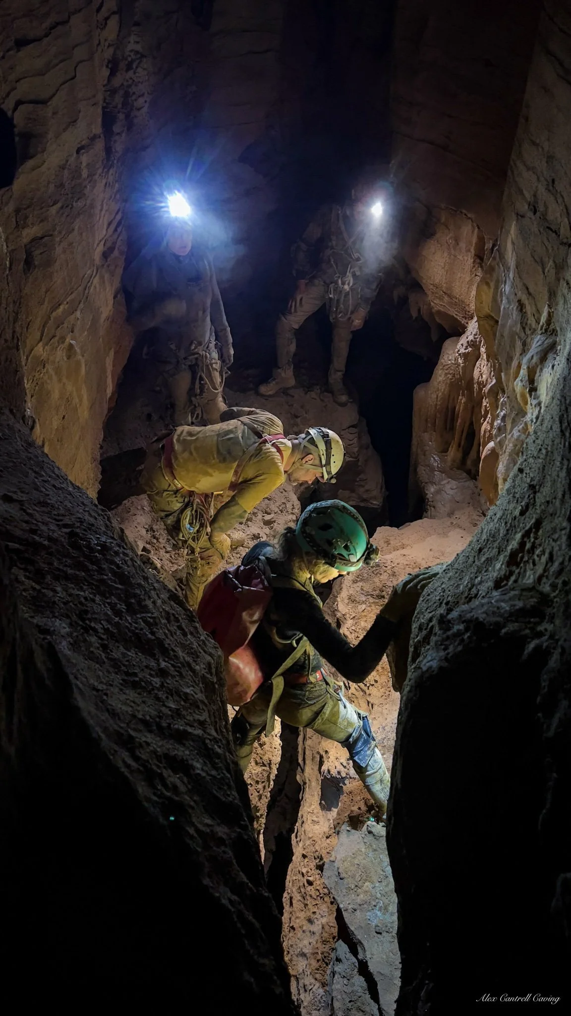Cave explorers wearing headlamps and protective gear navigating a narrow, rocky cavern.