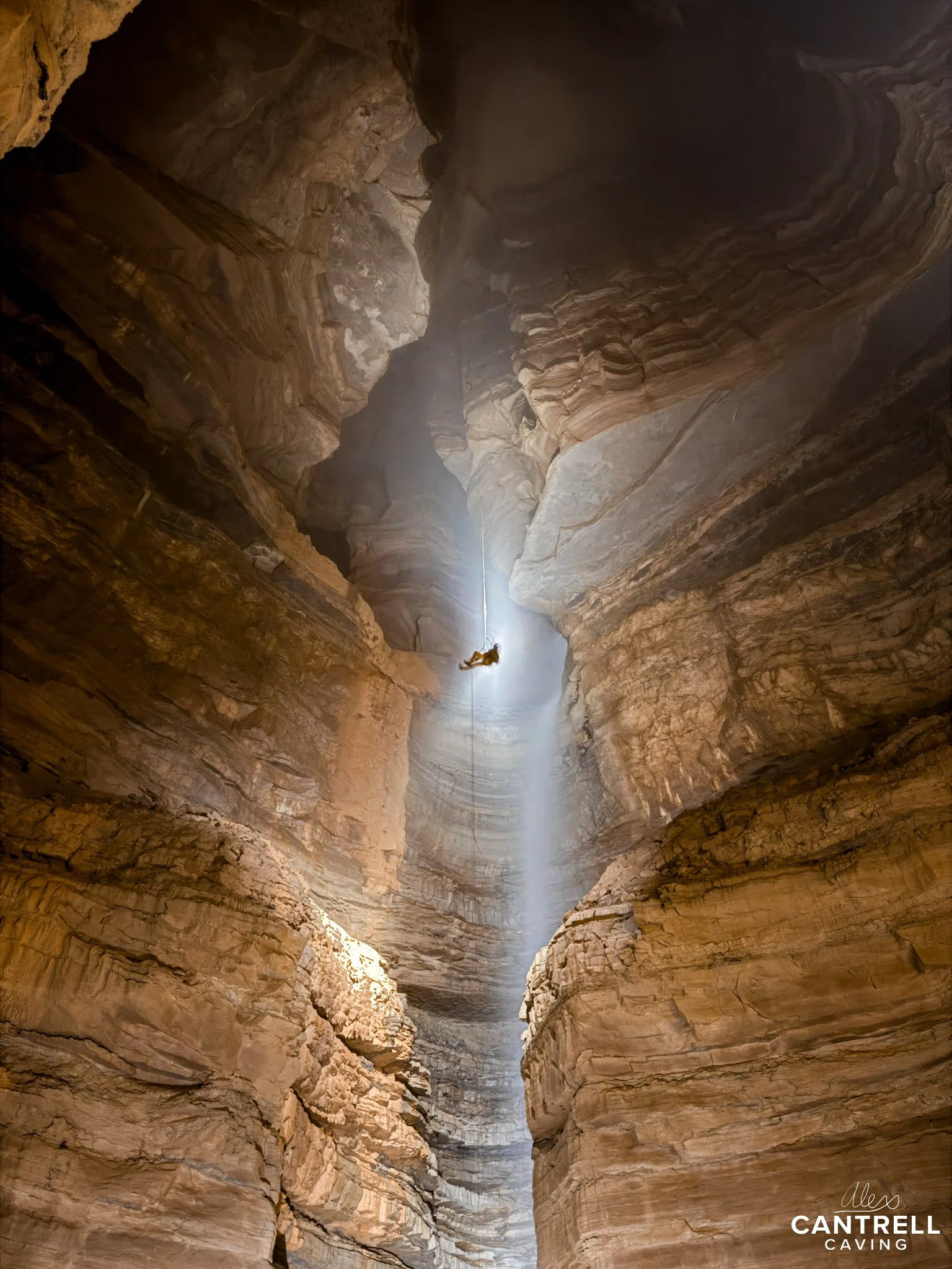 Caver descending into a large, illuminated cave shaft with a rope, surrounded by textured rock walls.