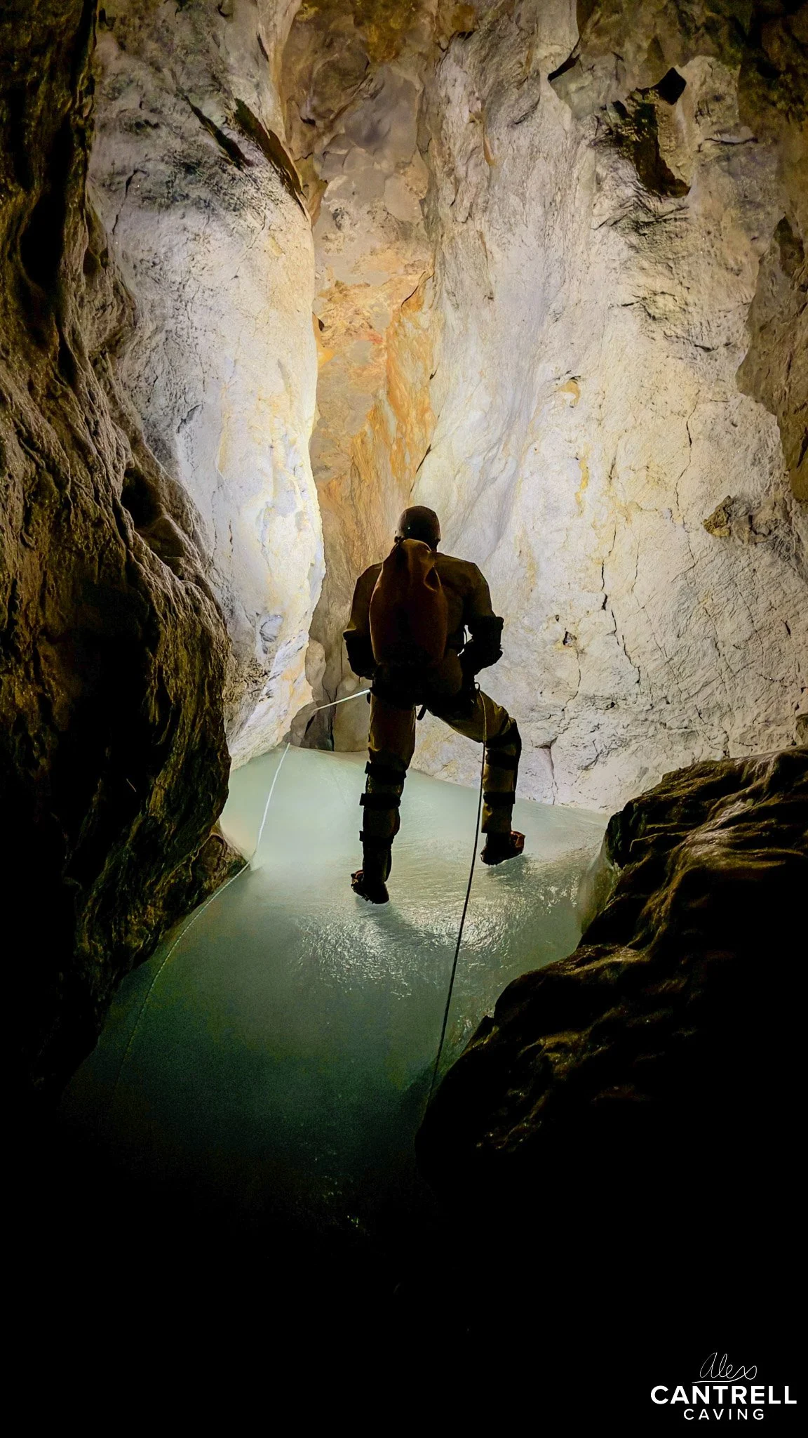 Person rappelling in cave with water below, wearing caving gear.