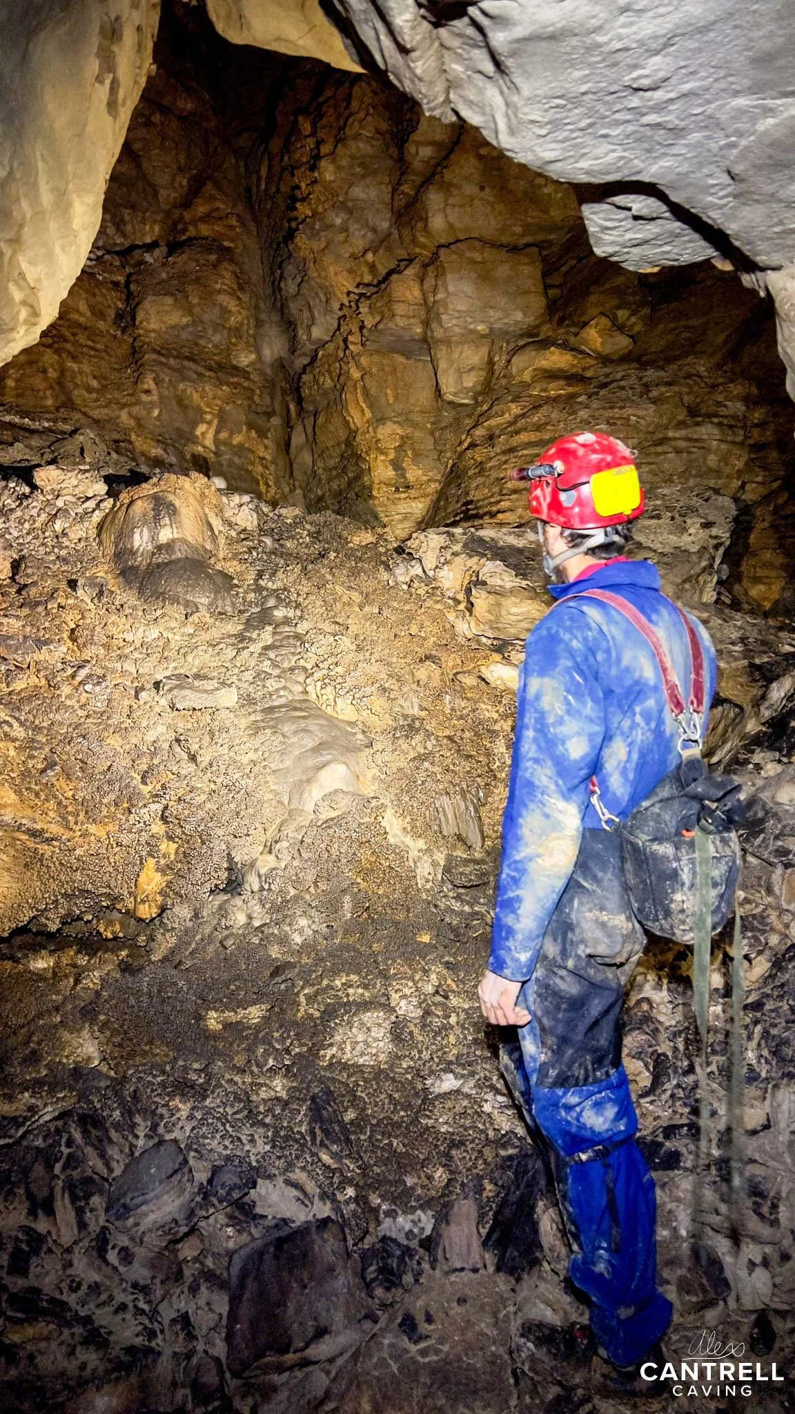 Caver exploring inside a cave wearing a red helmet and blue coveralls, surrounded by rocky walls and formations.