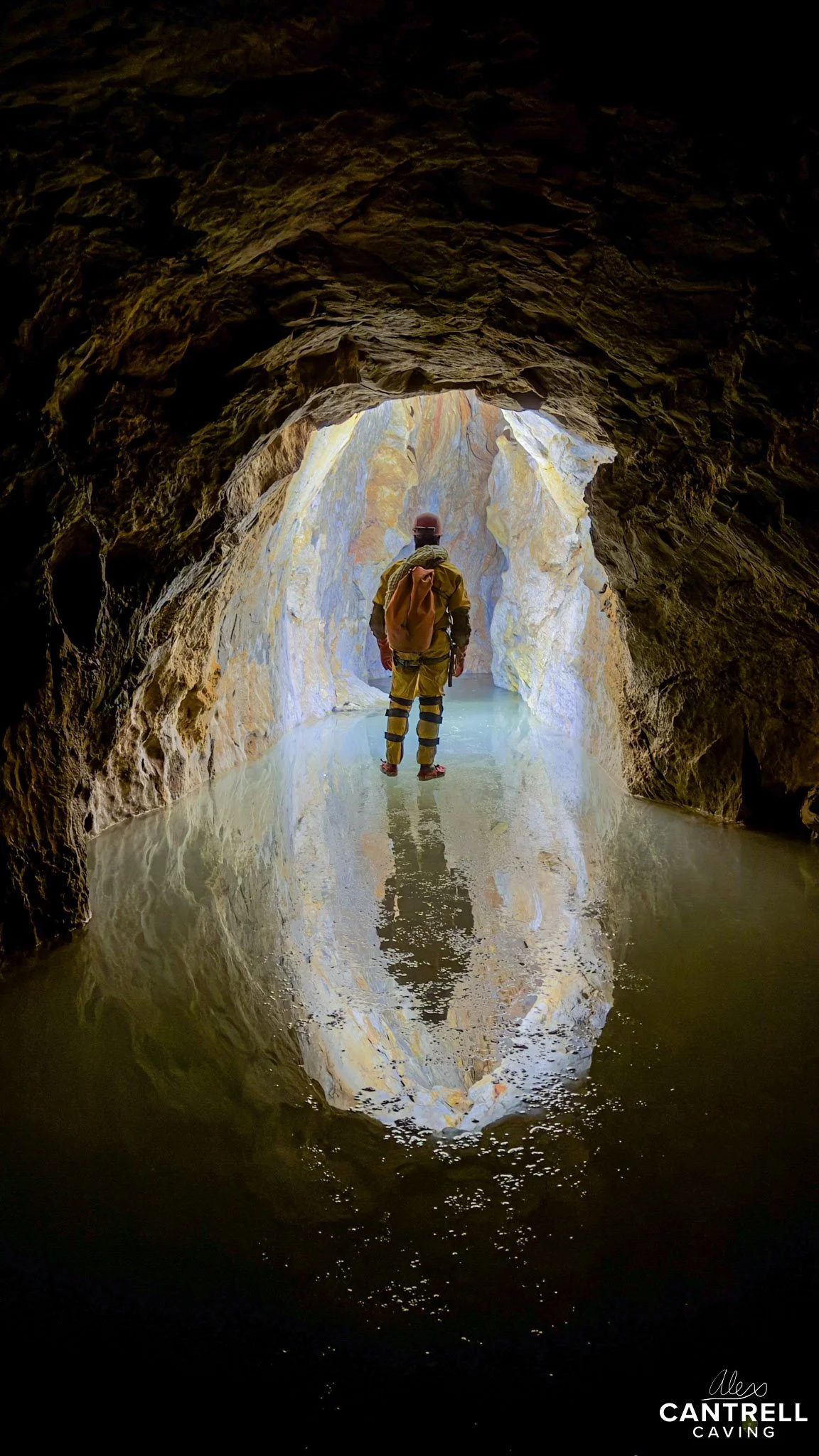 Person exploring a cave with water on the floor, wearing outdoor gear and carrying a backpack.