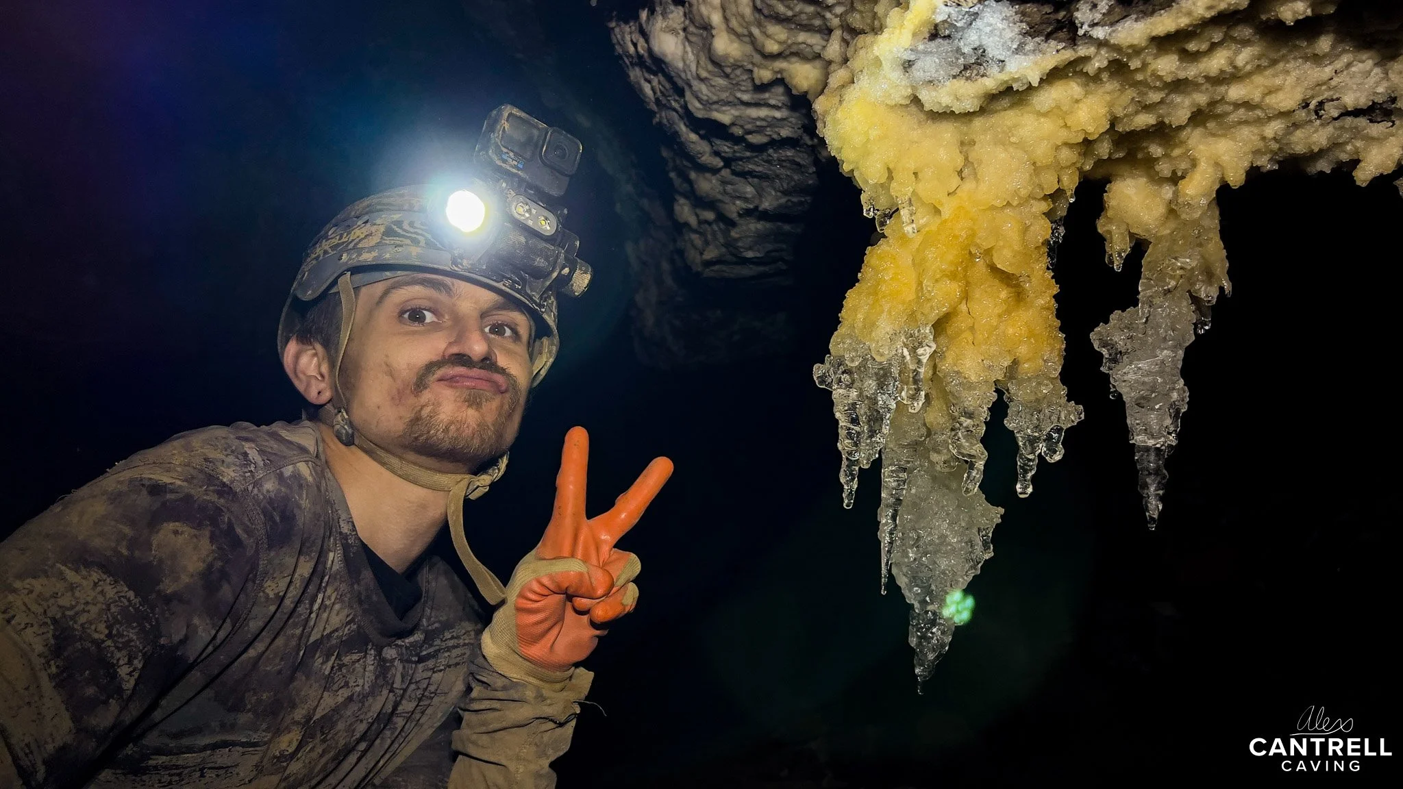 Caver posing in a cave with orange gloves, helmet, and headlamp next to stalactites.