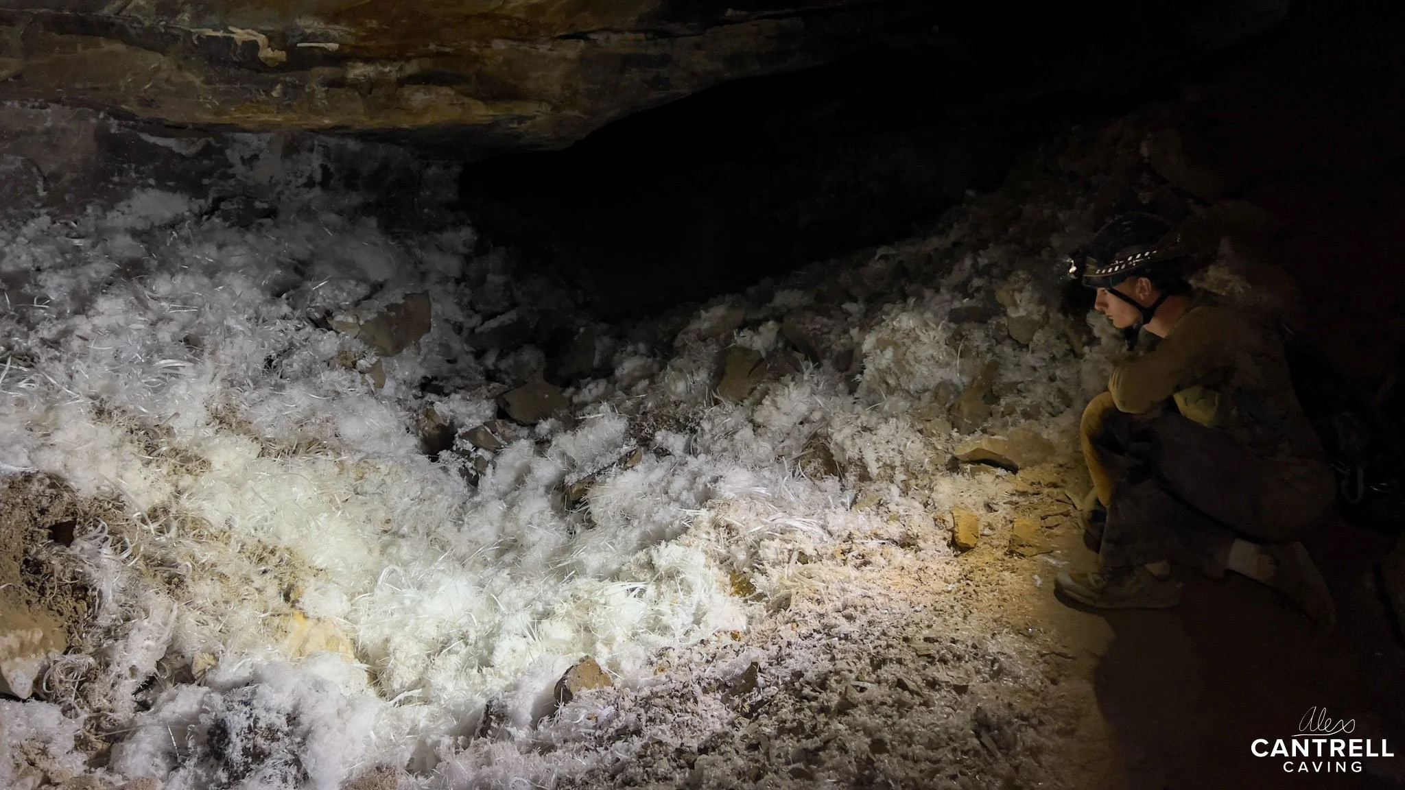 A person observing a cave formation with white crystals on the ground, under a rocky ceiling.