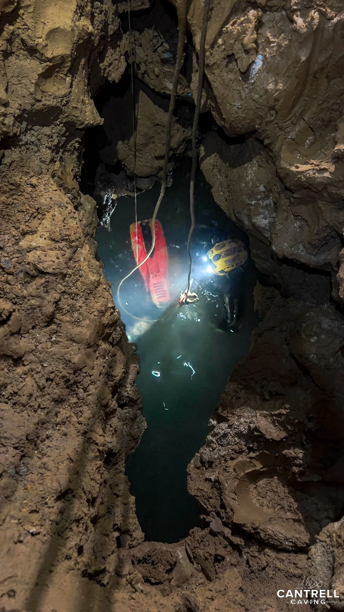 Underground cave with muddy walls, partially submerged equipment, water pool, ropes hanging, dim lighting