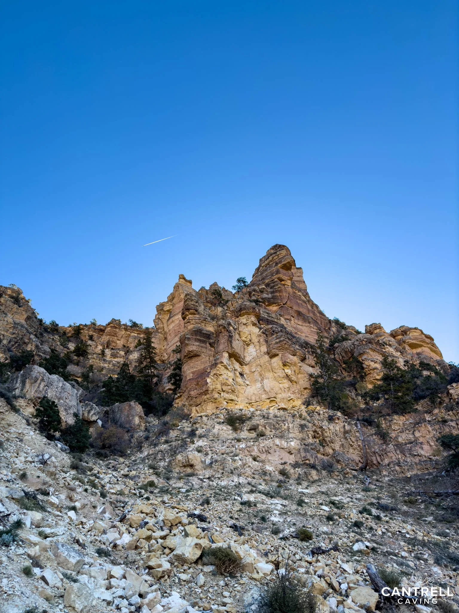 Towering cliff formation with rugged, rocky terrain and sparse vegetation under a clear blue sky. A contrail from an aircraft is visible in the sky.