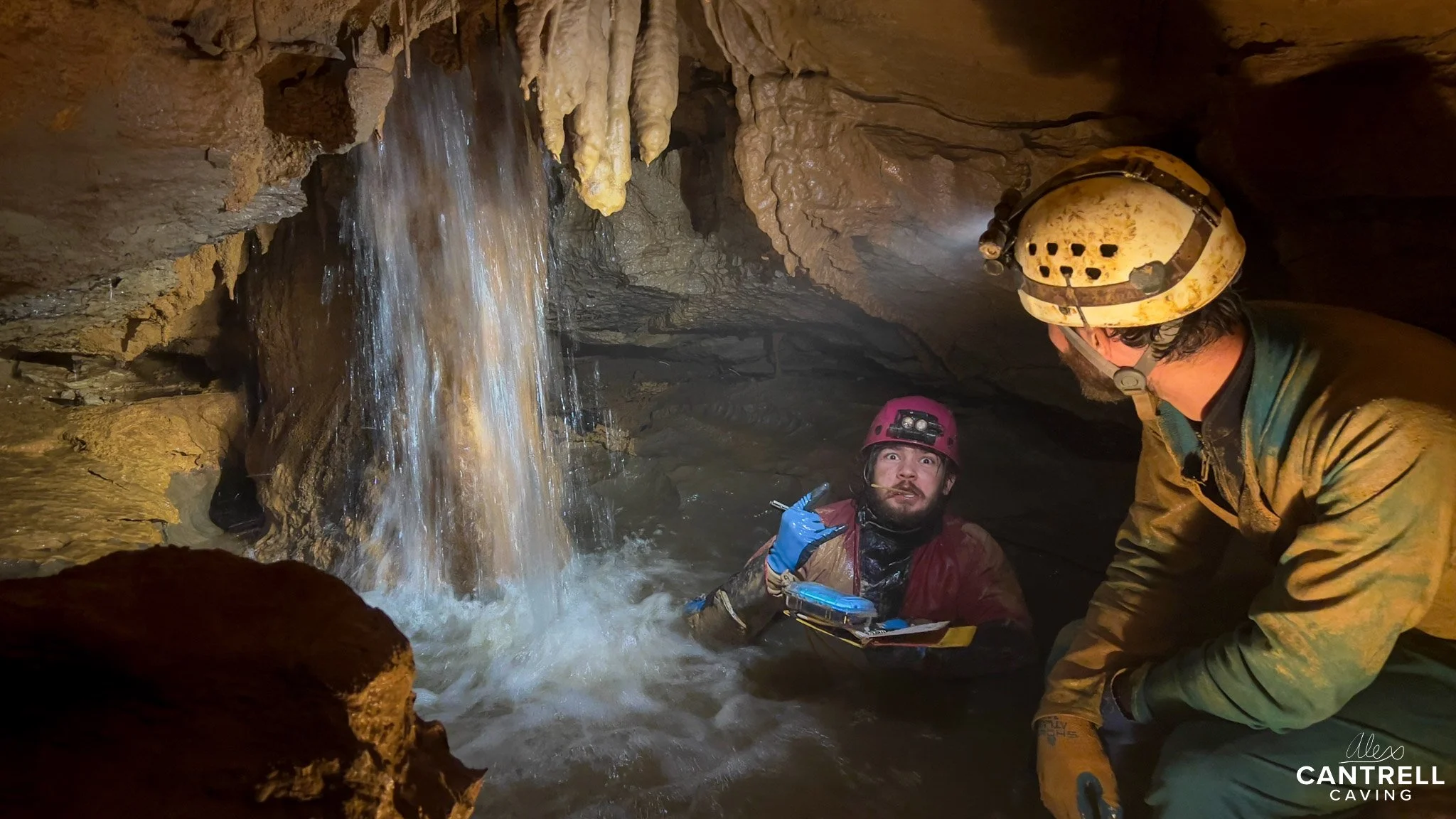 Two cavers in a cave with a waterfall, wearing helmets with lights and protective gear, surrounded by stalactites and cave formations.