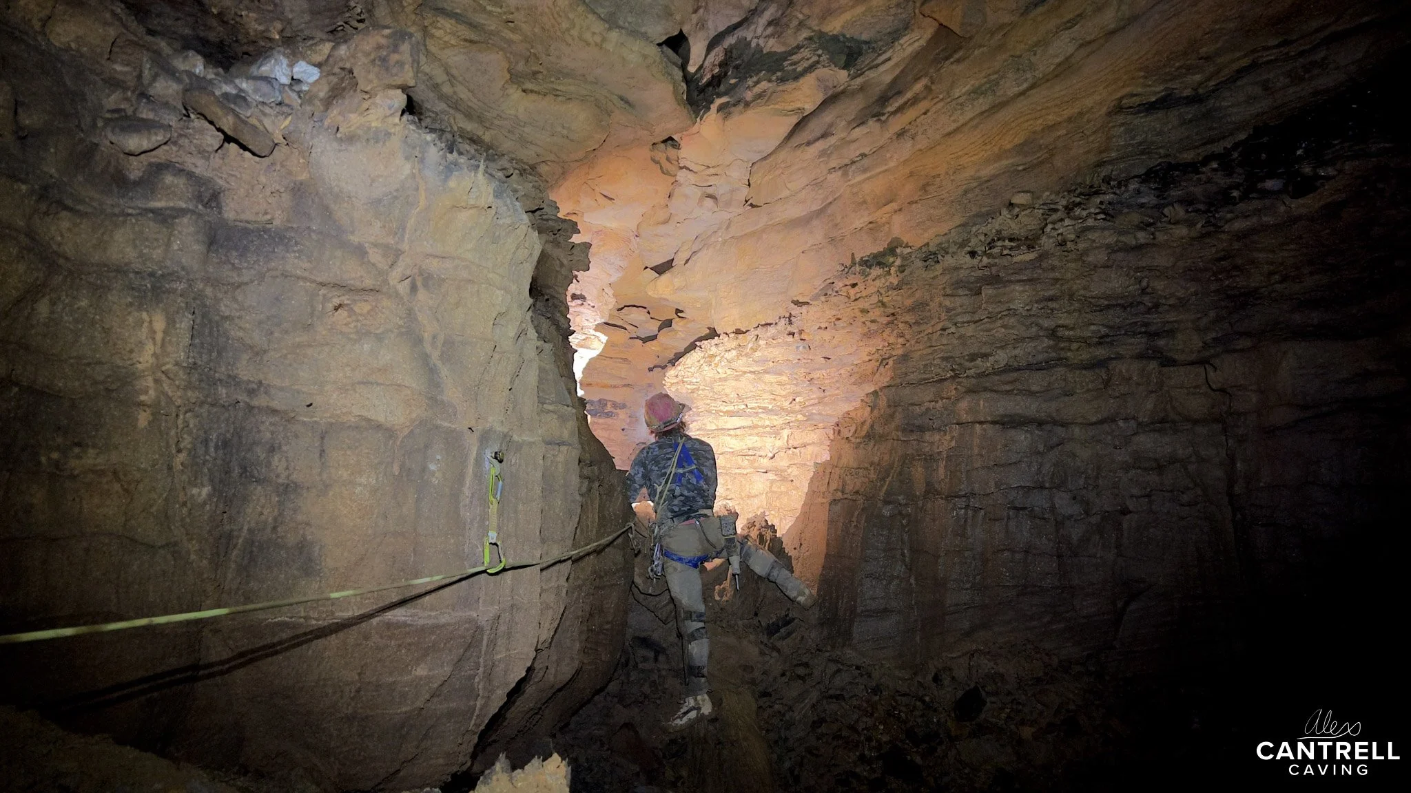 A person caving in a narrow passageway of a cave, using climbing gear and ropes for safety, with rugged rock formations and dim light.