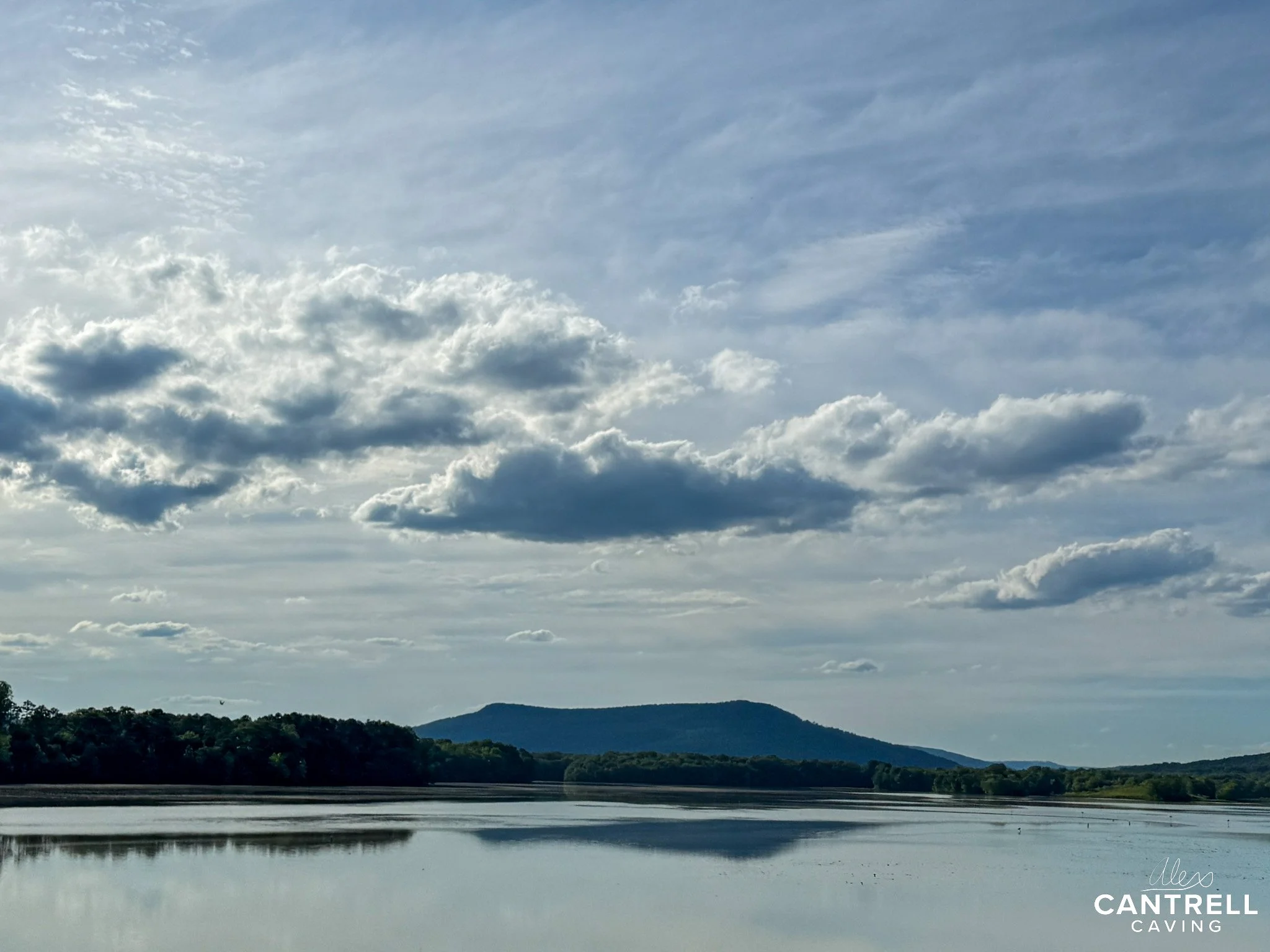 Scenic landscape of a calm lake with a reflection of the cloudy sky above, surrounded by lush green trees and distant hills under a partly cloudy sky.