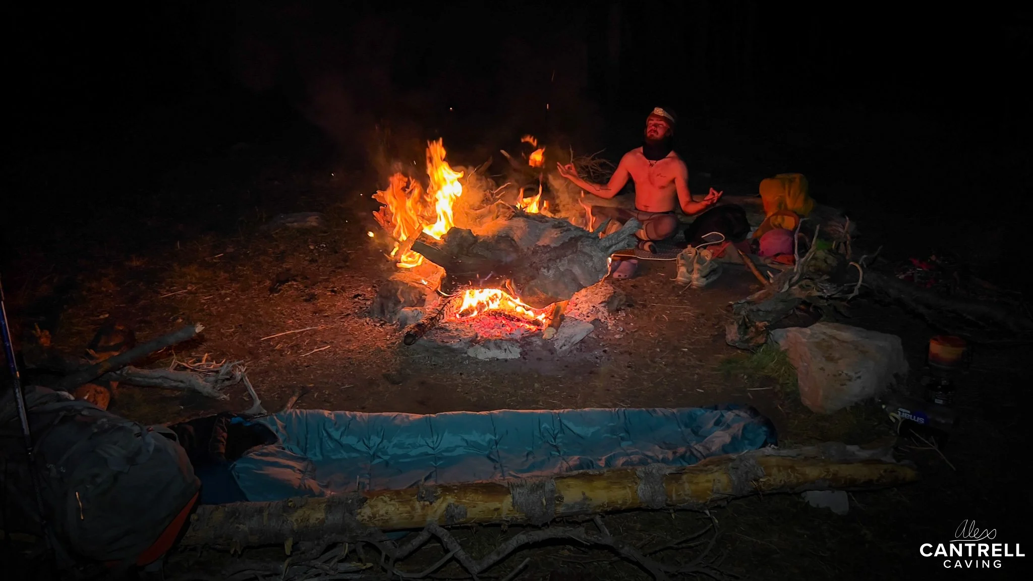 A person meditating in front of a campfire at night, surrounded by camping gear, with someone sleeping in a sleeping bag nearby.