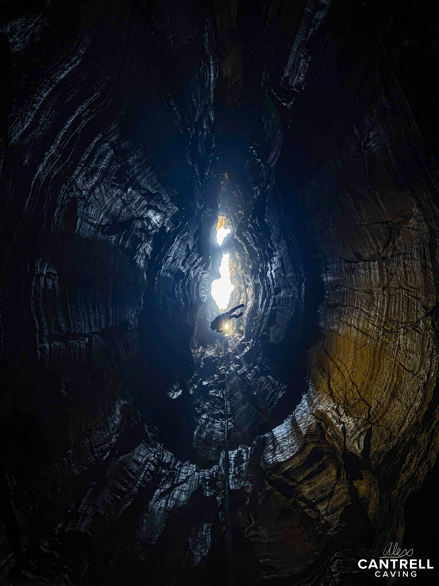 A climber descending into a deep, narrow cave with rugged rock walls, illuminated by a light source from above.
