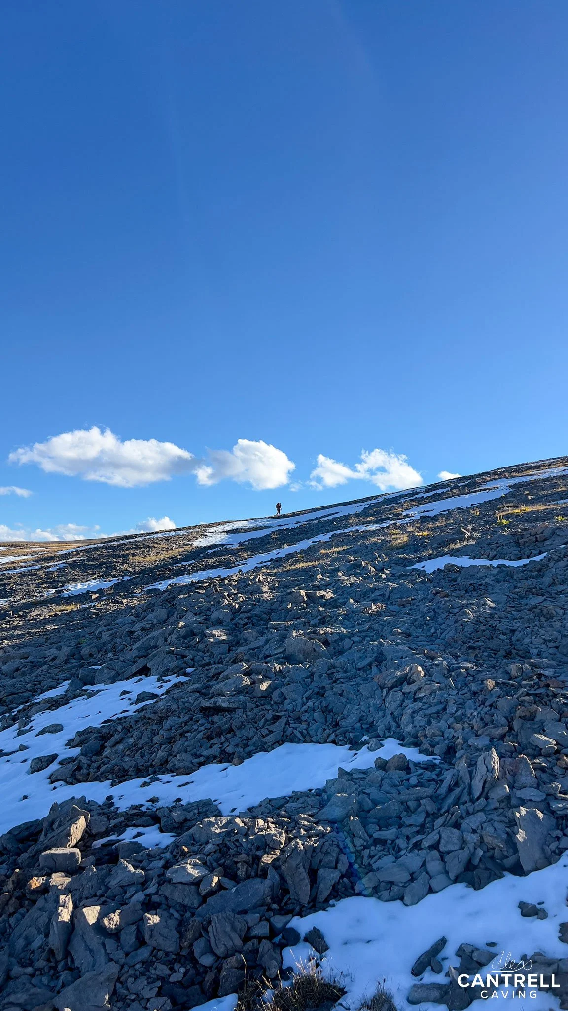 A person hiking on a rocky, snow-dusted hillside under a clear blue sky with scattered clouds.