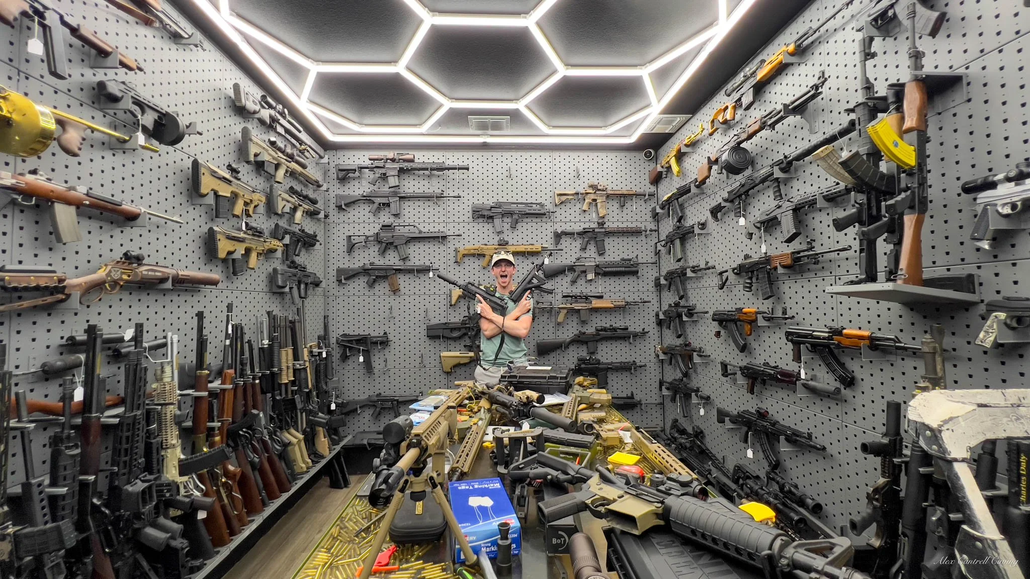 A man standing in a room filled with guns displayed on walls and a table. The room is well-lit with hexagonal lights on the ceiling. Various firearms, including rifles and shotguns, are mounted on pegboards on three walls. The table is cluttered with