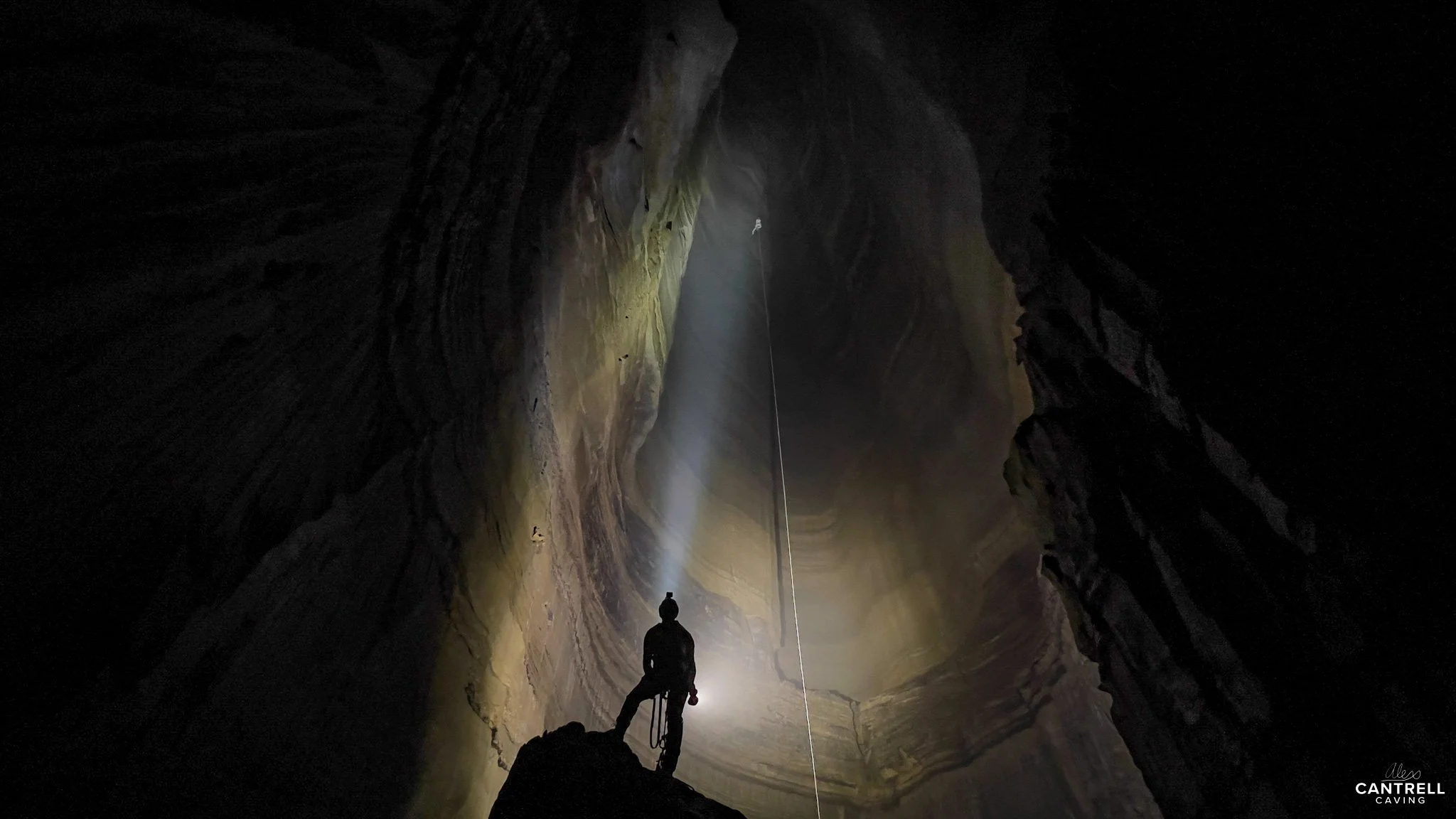 Silhouette of a caver standing at the entrance of a large, deep cave with a bright headlamp illuminating the stone walls.