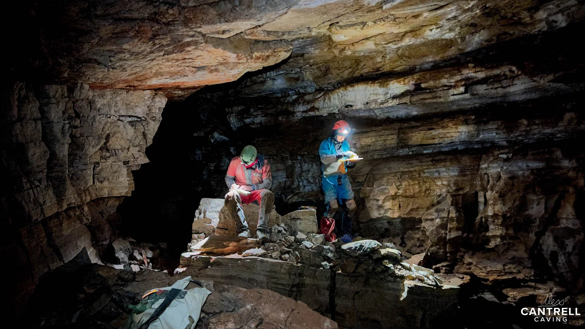 Two cavers with helmets and headlamps exploring a rocky cave, examining equipment or maps, surrounded by stone walls and formations.