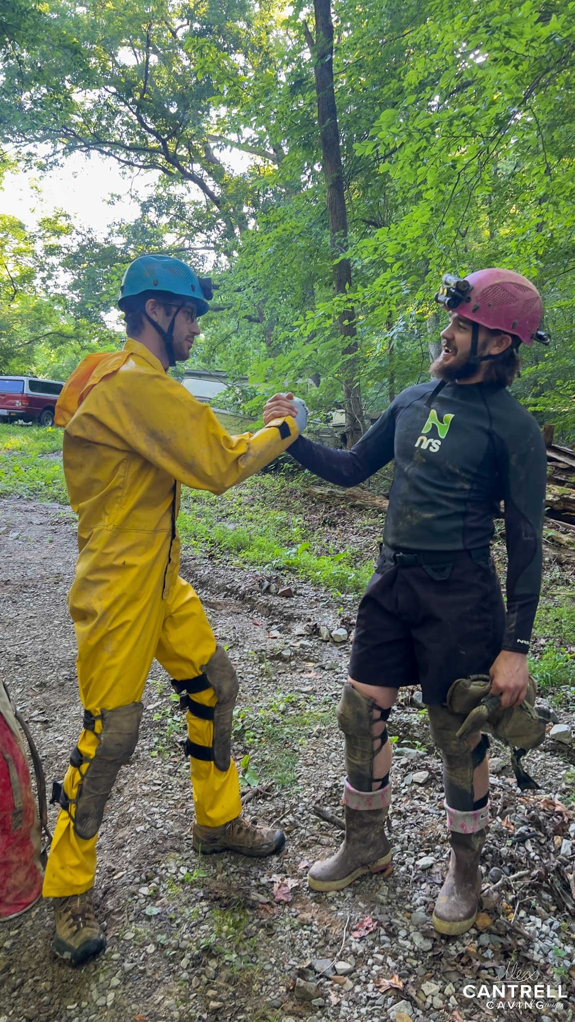 Two cavers in a forest wearing protective gear and helmets shake hands on a rocky path.