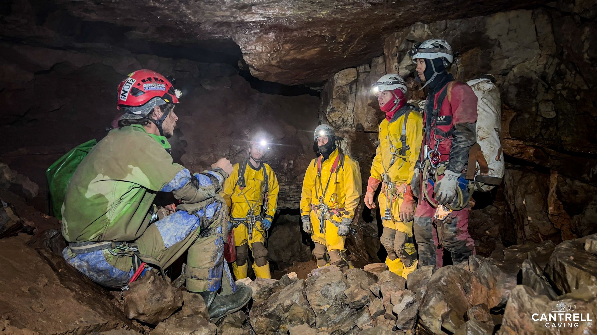 Group of cavers in a cave, wearing helmets and headlamps, standing on rocky ground.
